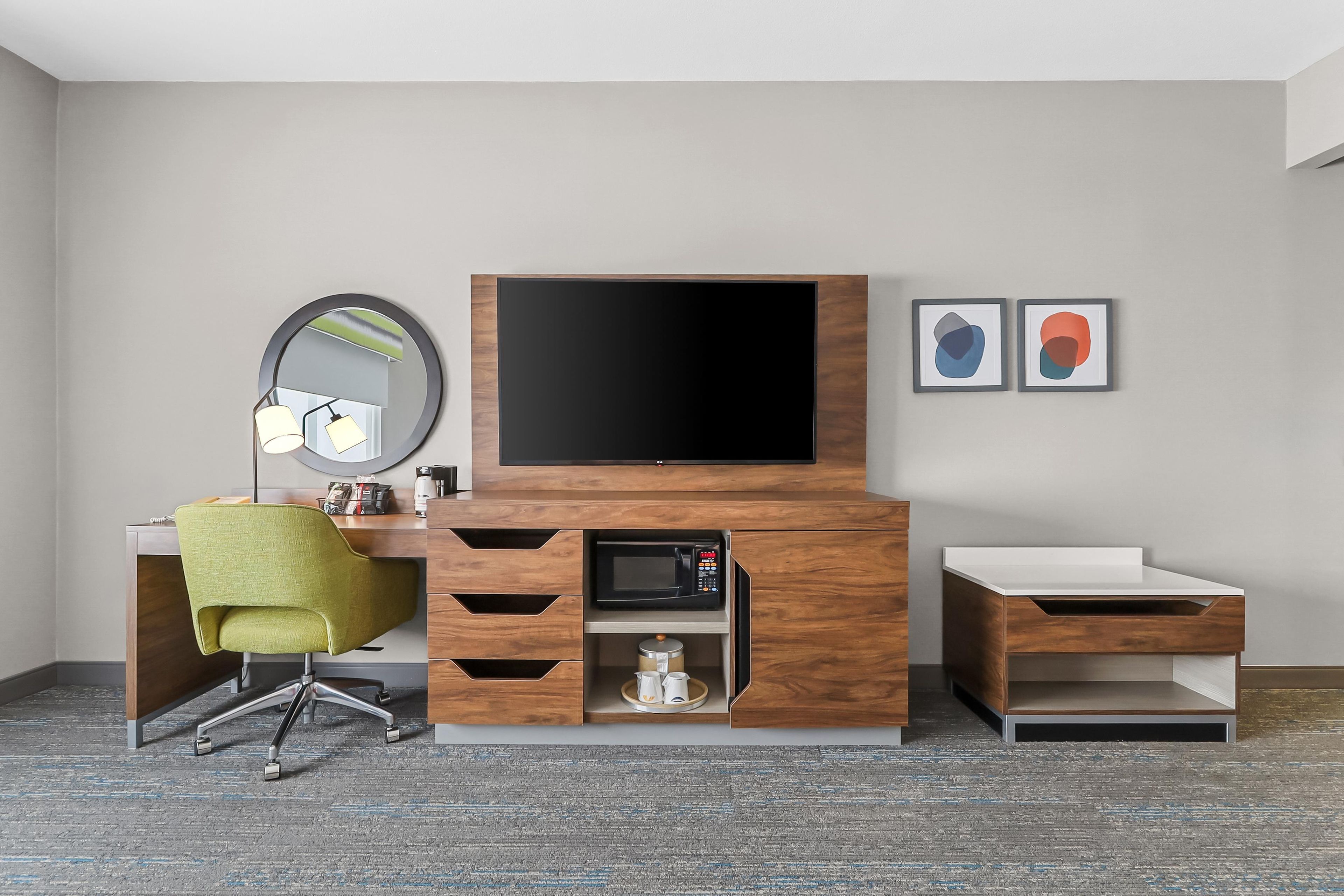 A modern hotel room with a wooden desk, a flat-screen TV, a mirror, and two framed artworks.