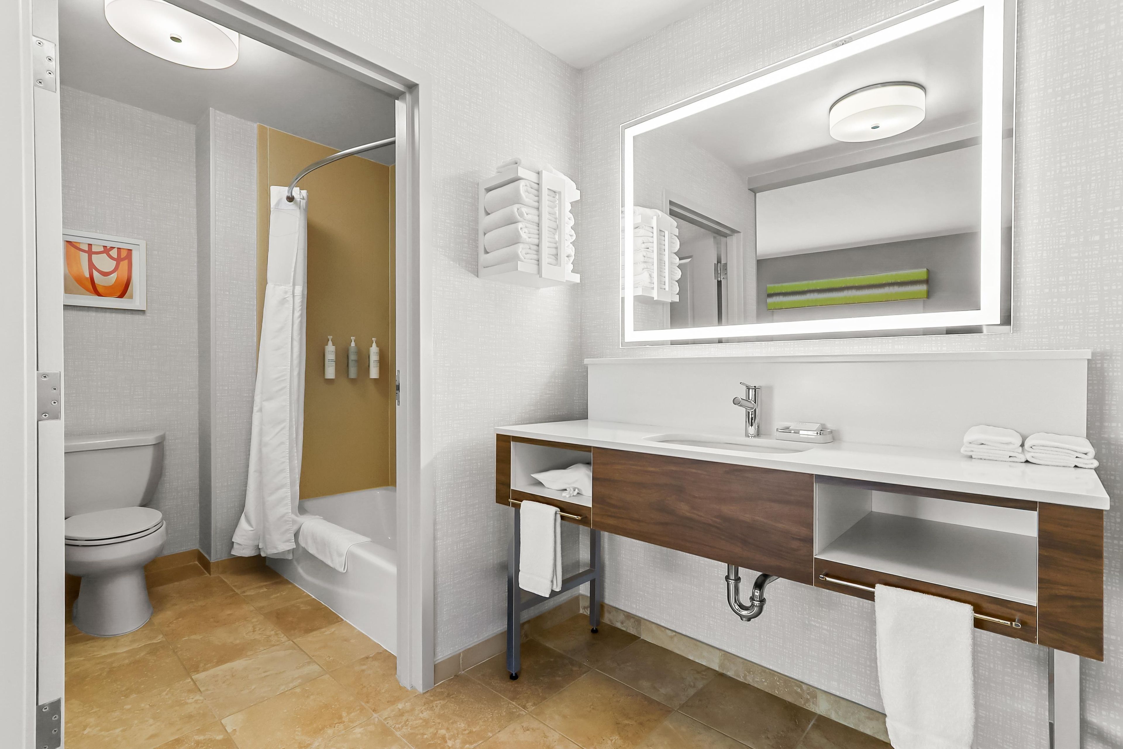 A modern bathroom with a toilet, shower, and sink, featuring a mirror, towel rack, and a framed picture on the wall.