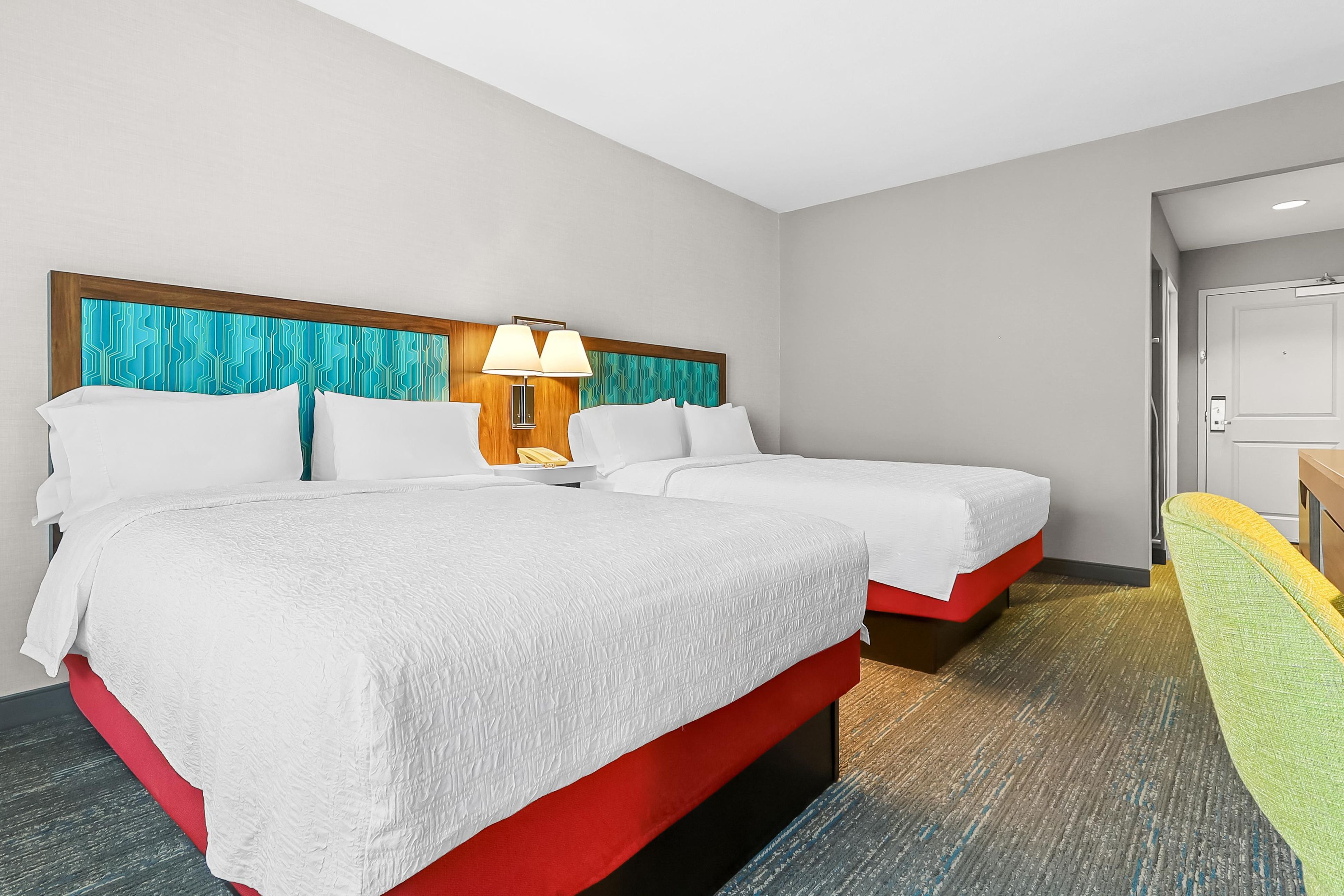 Ein Hotelzimmer mit zwei Betten, einem Nachttisch mit Lampe, einem Holz-Kopfteil und einer Tür rechts.