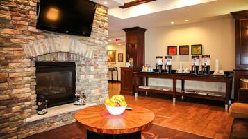 A stone fireplace, a television mounted above it, a coffee station with multiple dispensers, and a bowl of fruit on a wooden table.