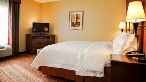 A hotel room with a bed, a television on a wooden cabinet, a framed picture on the wall, and a desk with a lamp and phone.