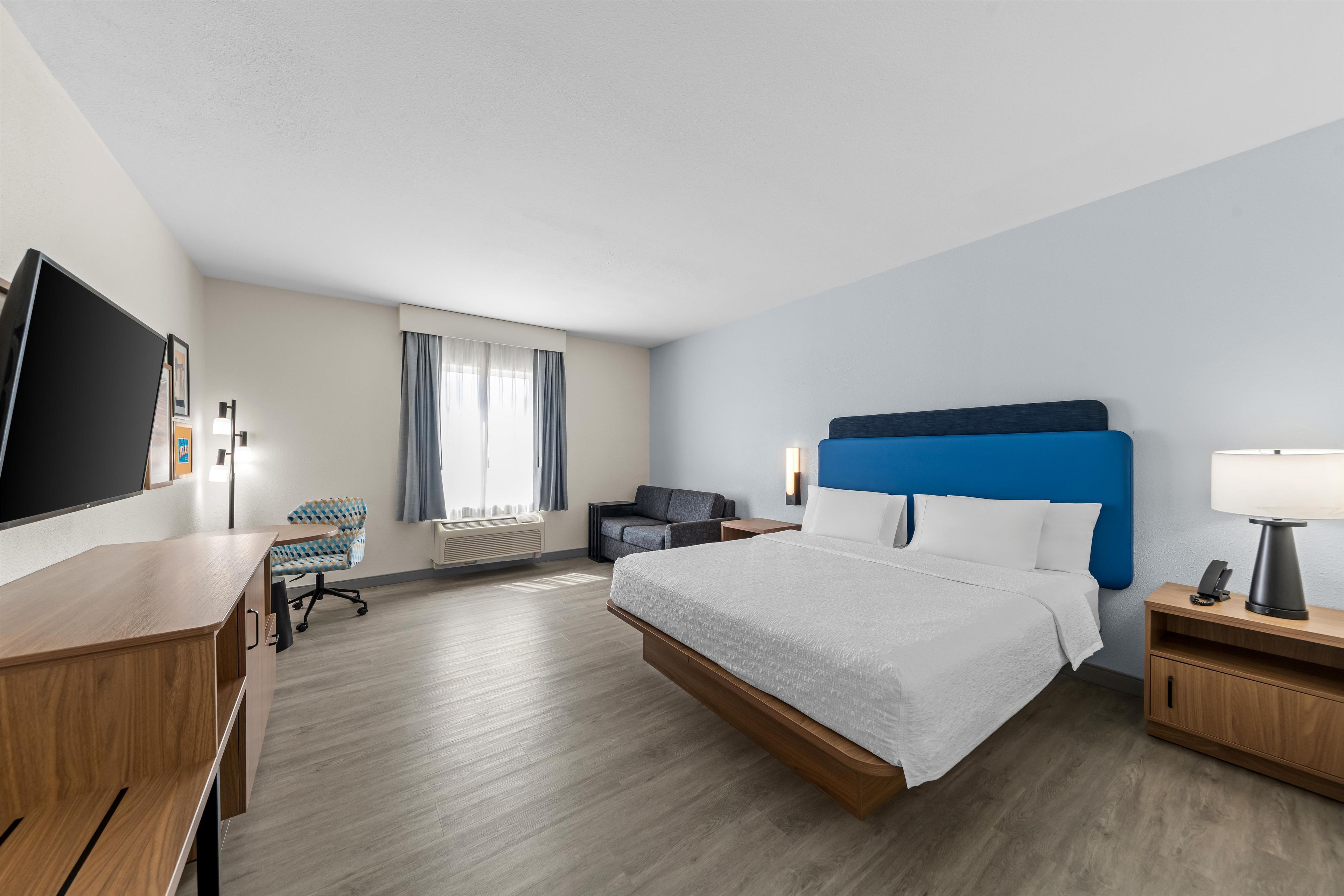 Quarto de hotel com cama, escrivaninha, cadeira e televisão.