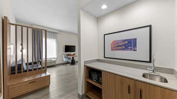 Una habitación de hotel moderna con un armario de madera, un televisor de pantalla plana, un cuadro enmarcado con el cartel de un ferrocarril estatal de Texas y una zona de cocina con lavamanos y microondas.