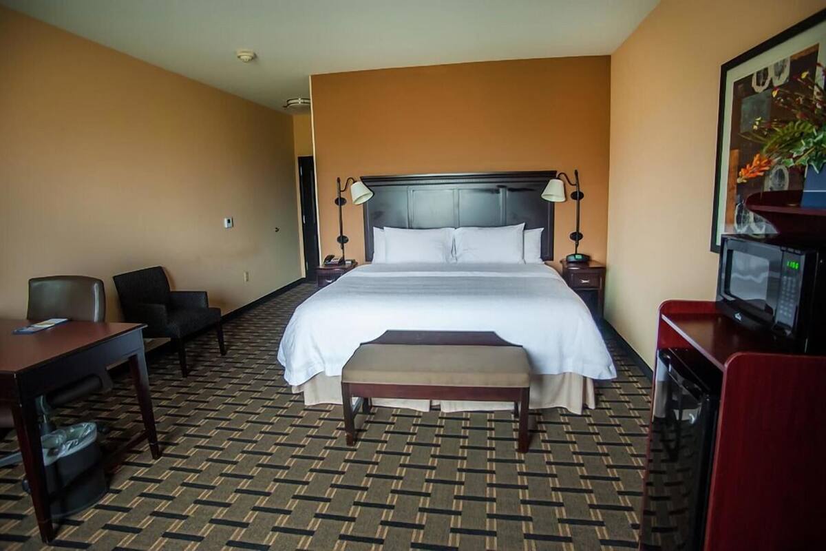 A hotel room with a large bed, a desk, a chair, a bench, a TV, and a microwave.