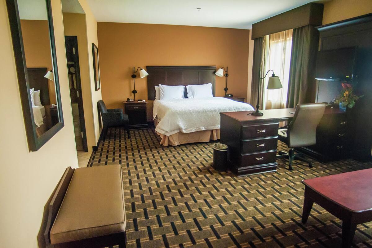A hotel room with a bed, a desk, a chair, a dresser, and a bench.