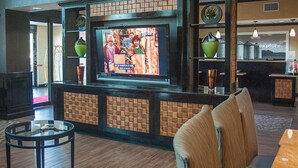 A lobby with a television mounted on a brick wall, seating area with a round table, and a reception desk in the background. - Hampton Inn & Suites Palestine (Palestine)