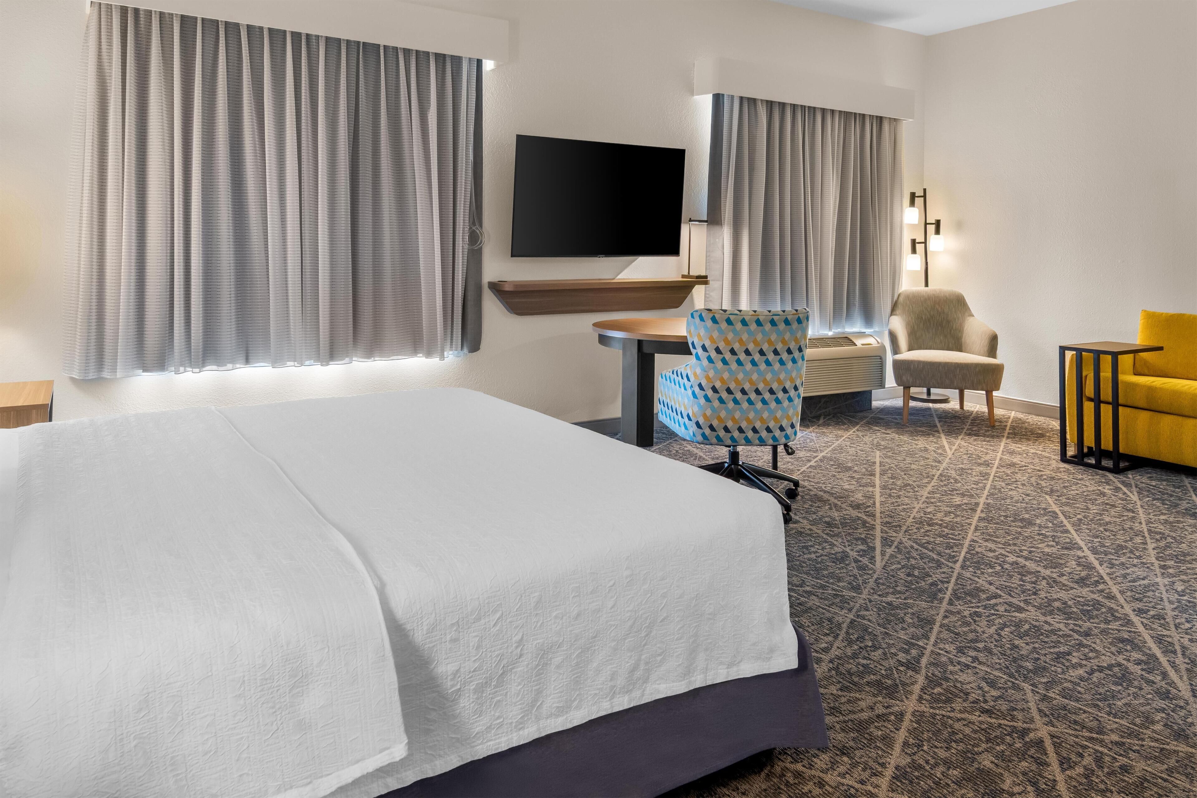 Una habitación de hotel con cama, escritorio, televisor, dos sillas y lámpara.