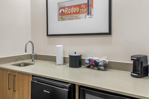 Una cocina de hotel con un lavamanos, dispensador de toallas de papel, cafetera y un cubo de basura.