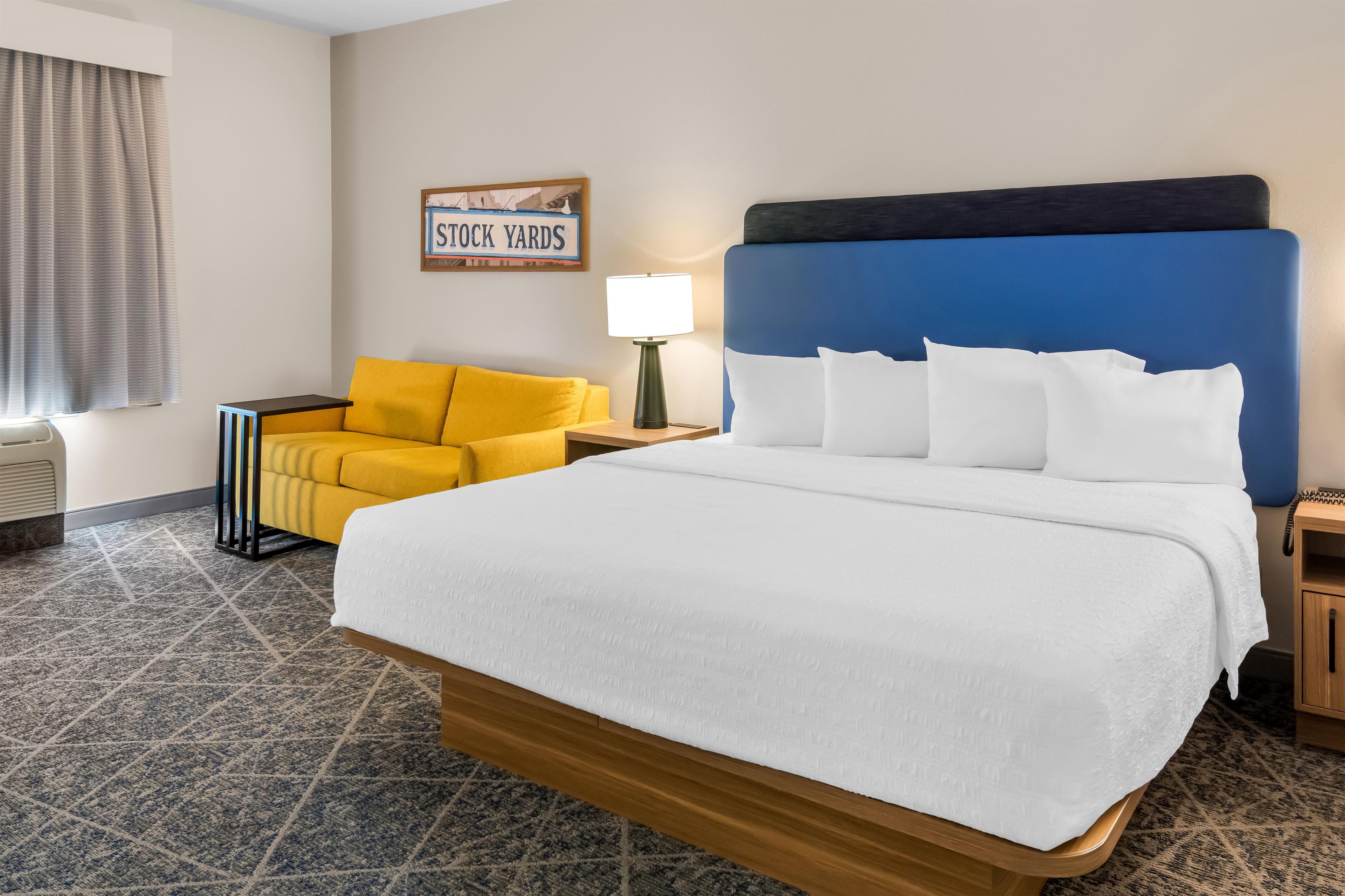 A hotel room with a large bed, a yellow armchair, a nightstand with a lamp, and a sign on the wall.