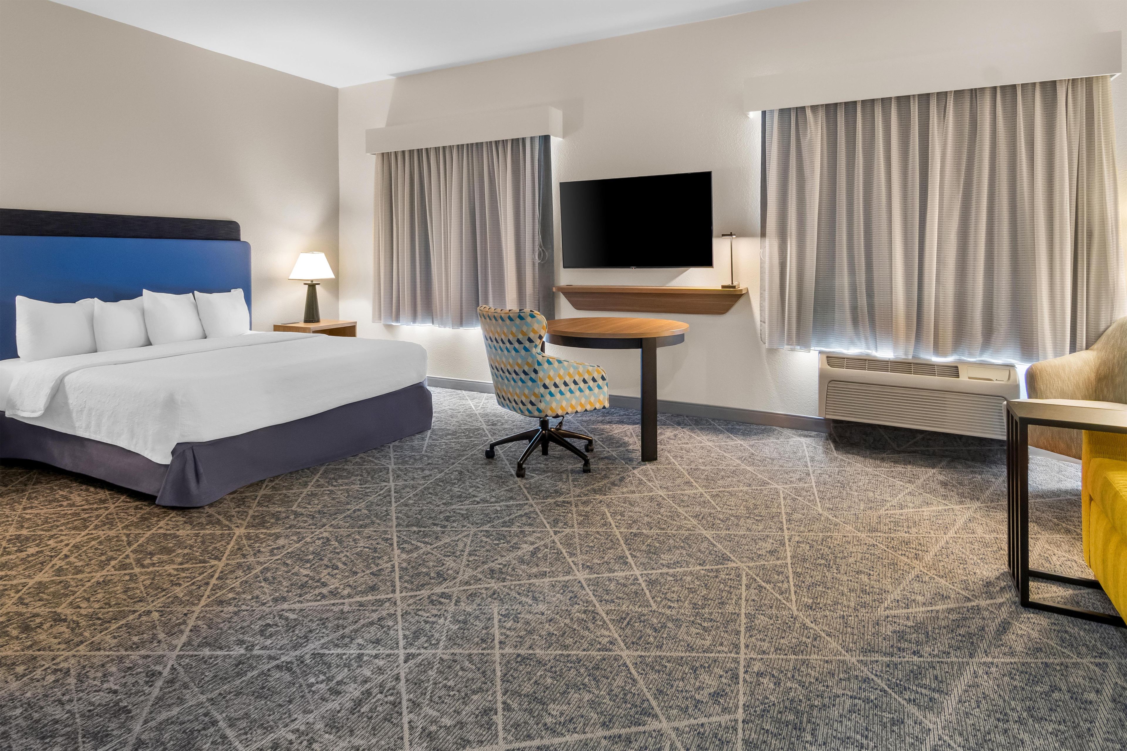 Quarto de hotel com cama, escrivaninha, cadeira, televisão de tela plana e janela com cortinas.