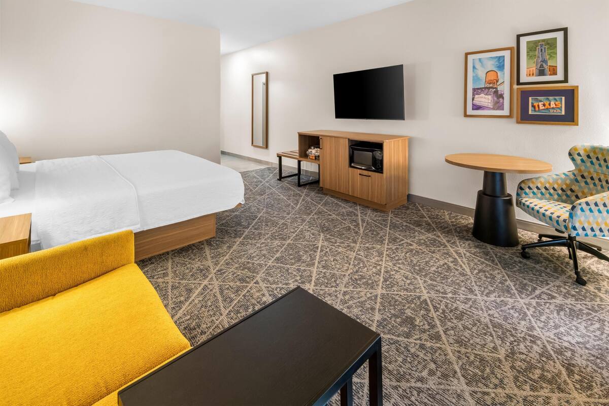 A hotel room with a bed, a sofa, a small table, a TV, a microwave, and framed pictures on the wall.