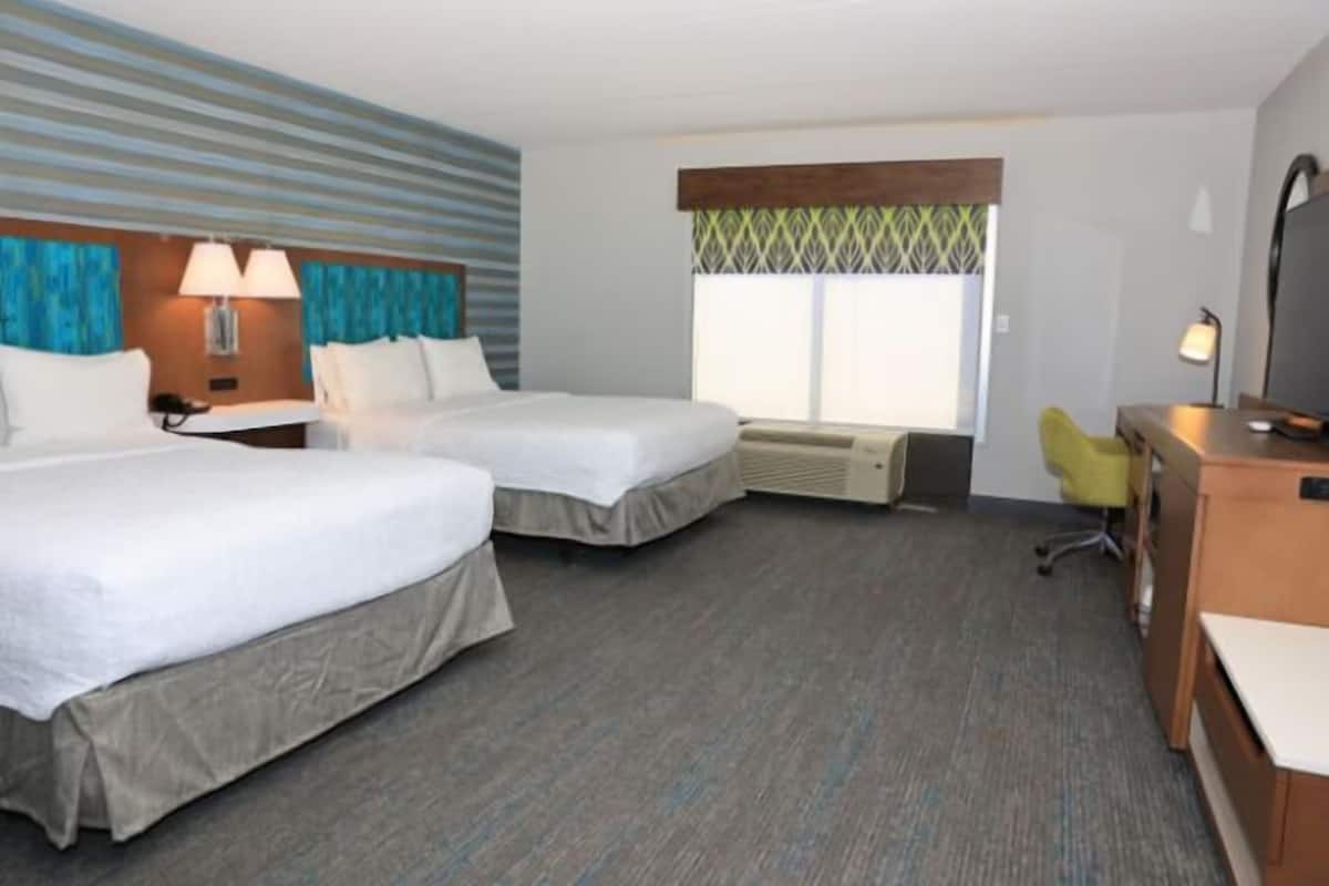 Quarto de hotel com duas camas, uma escrivaninha, uma cadeira, uma televisão e uma janela com vista.