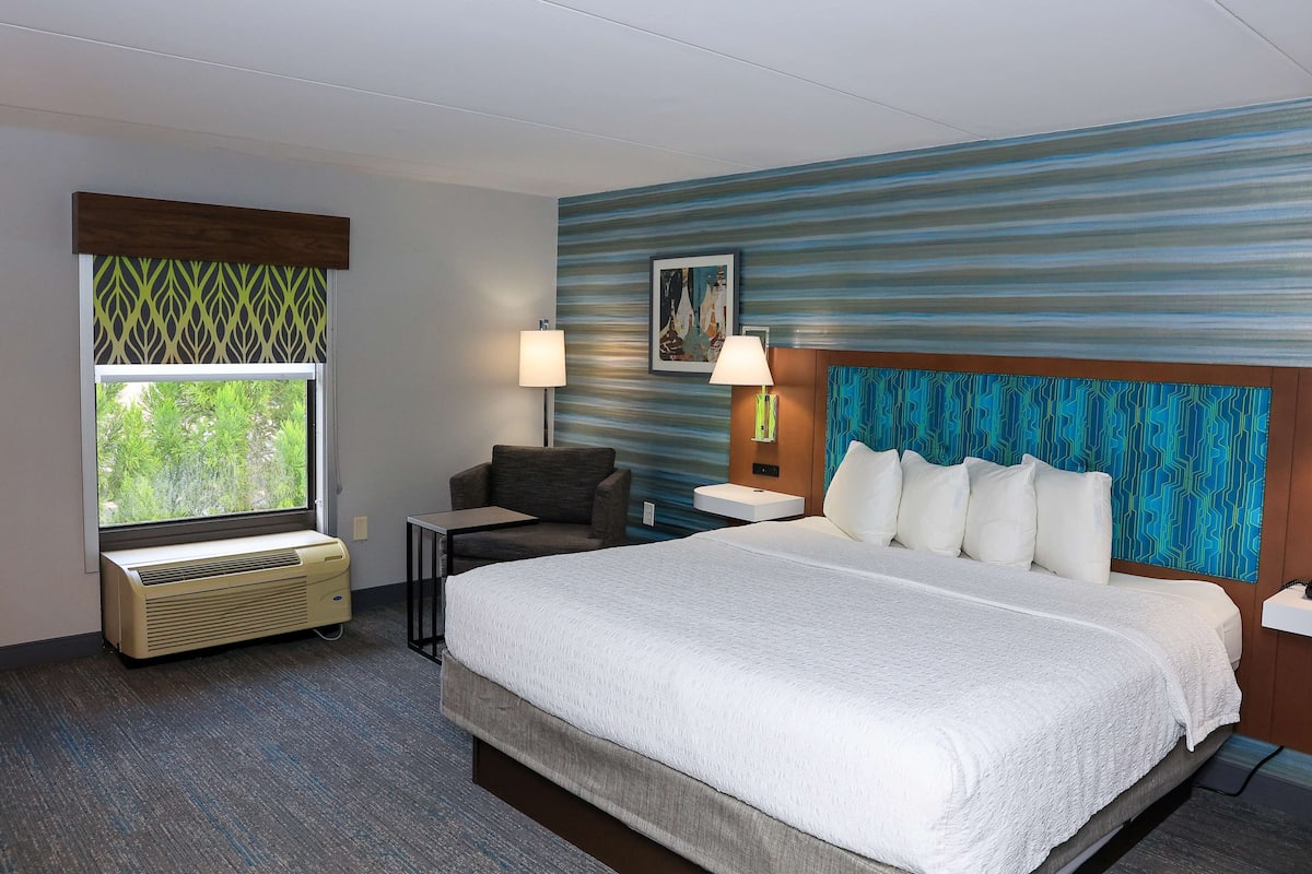 Quarto de hotel com uma cama grande, uma mesa, uma cadeira, uma janela com vista e uma parede com padrão azul e verde.