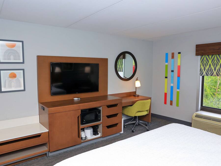 A hotel room with a bed, a desk with a chair, a television, a microwave, a coffee maker, and a window with a view of greenery.