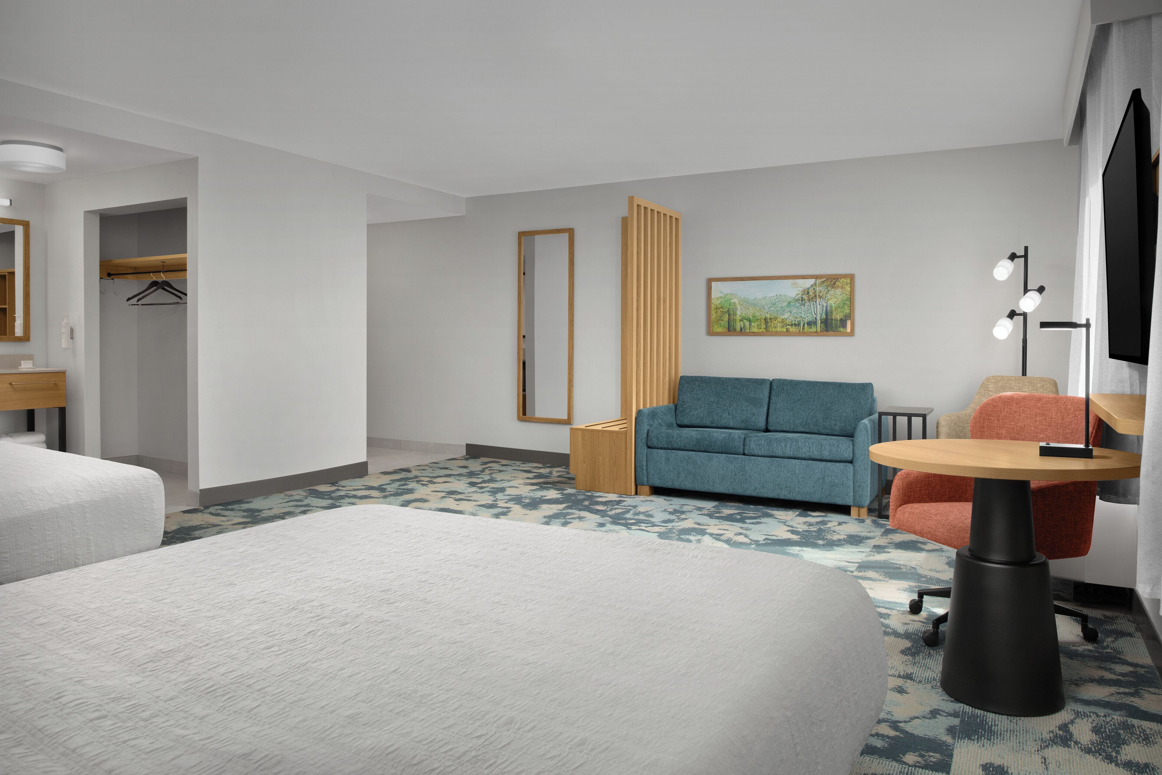 A hotel room with a large bed, a sofa, a small table, and a desk. There is a wardrobe, a painting on the wall, and a lamp.