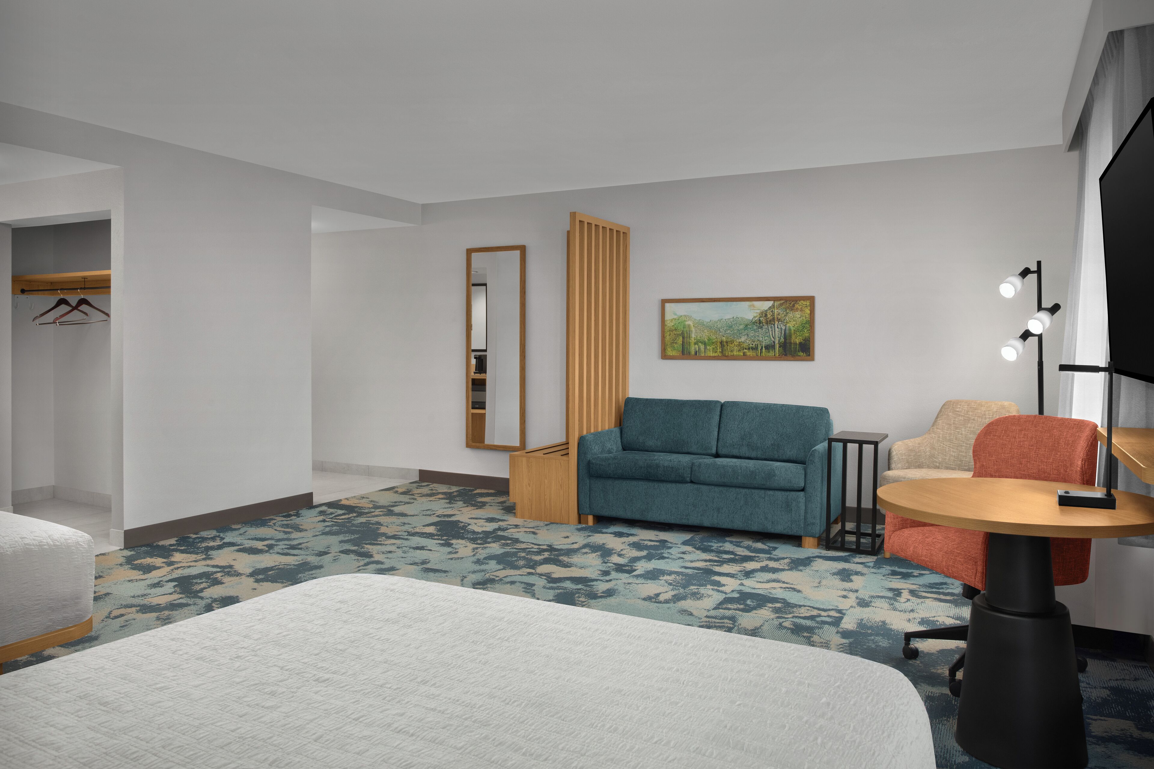 A hotel room with a bed, a sofa, a chair, a small table, a TV, a painting, and a closet.