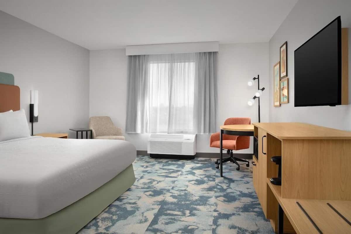 Quarto de hotel com cama, secretária, cadeira, janela com cortinas, televisão na parede e uma mesa pequena.