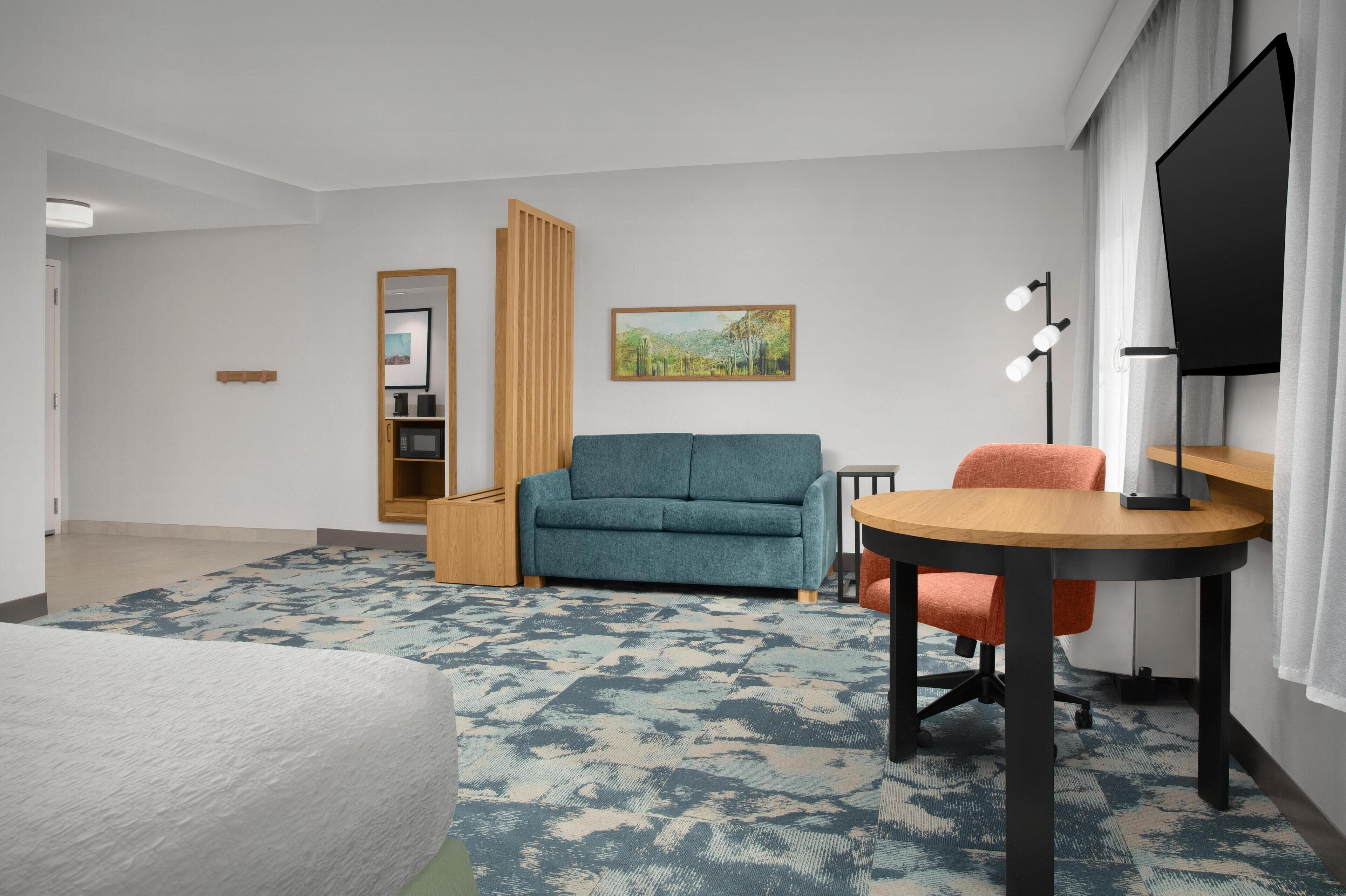 A hotel room with a sofa, a small table, a TV, a lamp, a bookshelf, and a painting on the wall.