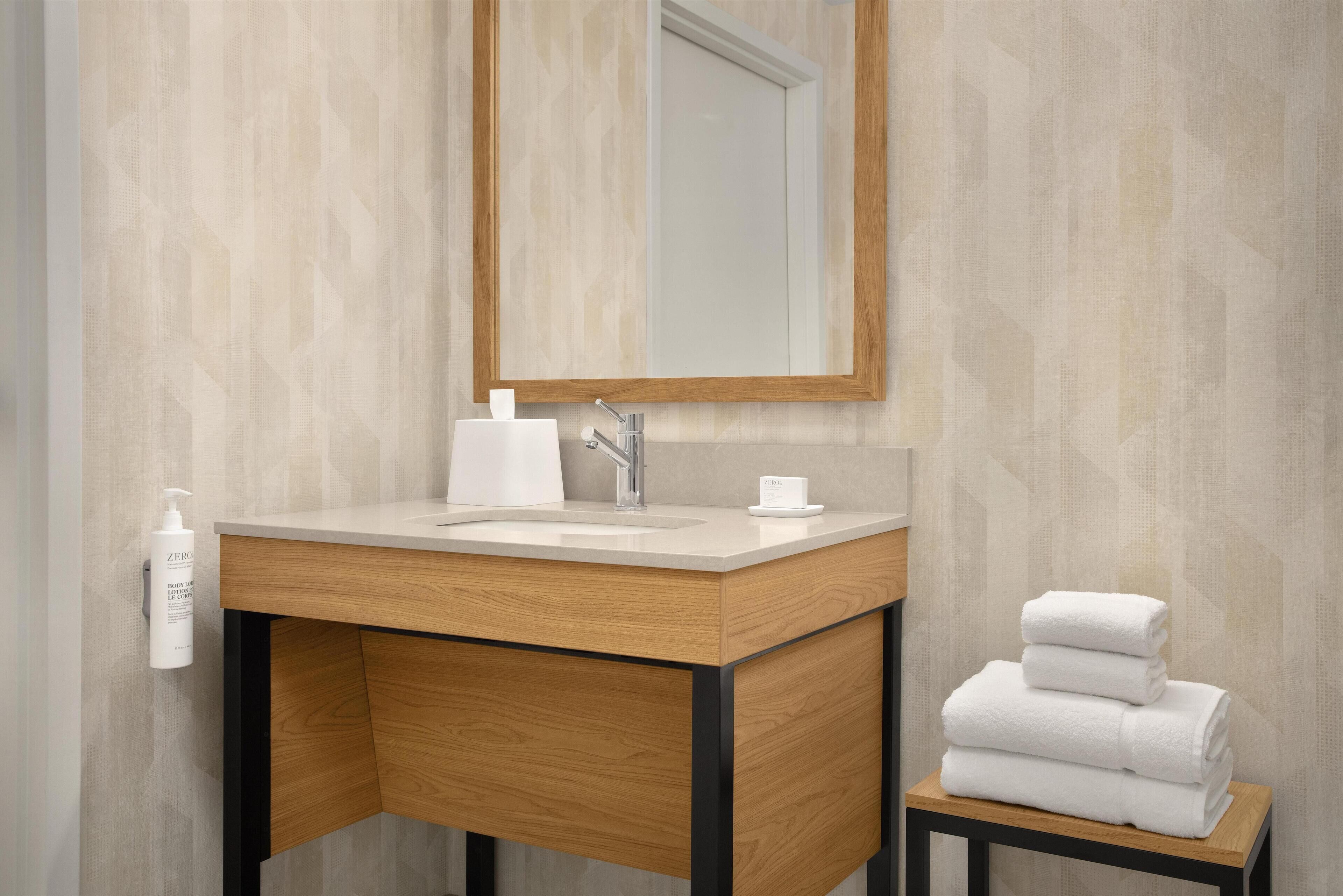 A modern bathroom with a wooden vanity, a sink, a mirror, and a stack of white towels on a small table.