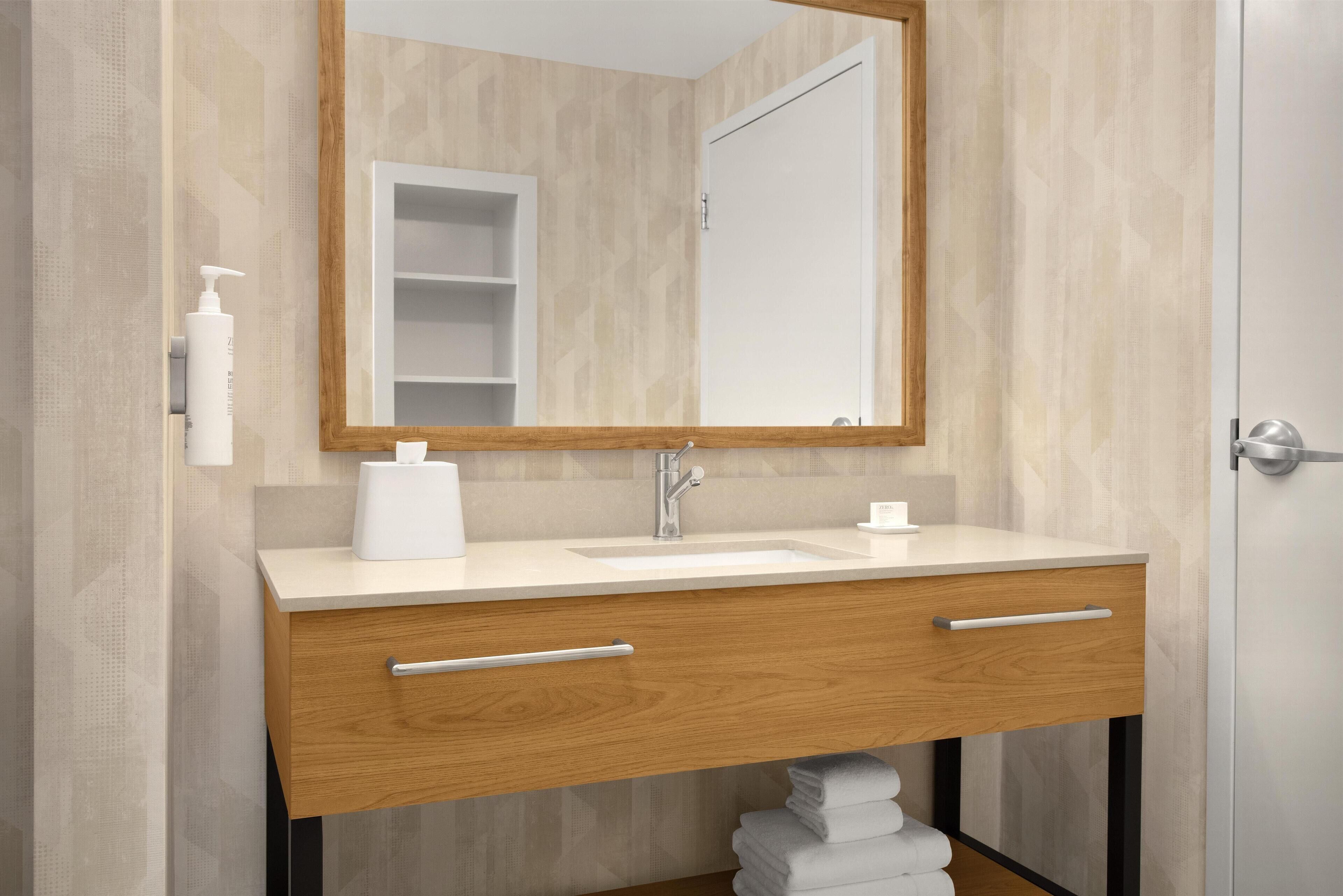 A bathroom with a wooden vanity, a white sink, a mirror, and a towel dispenser.