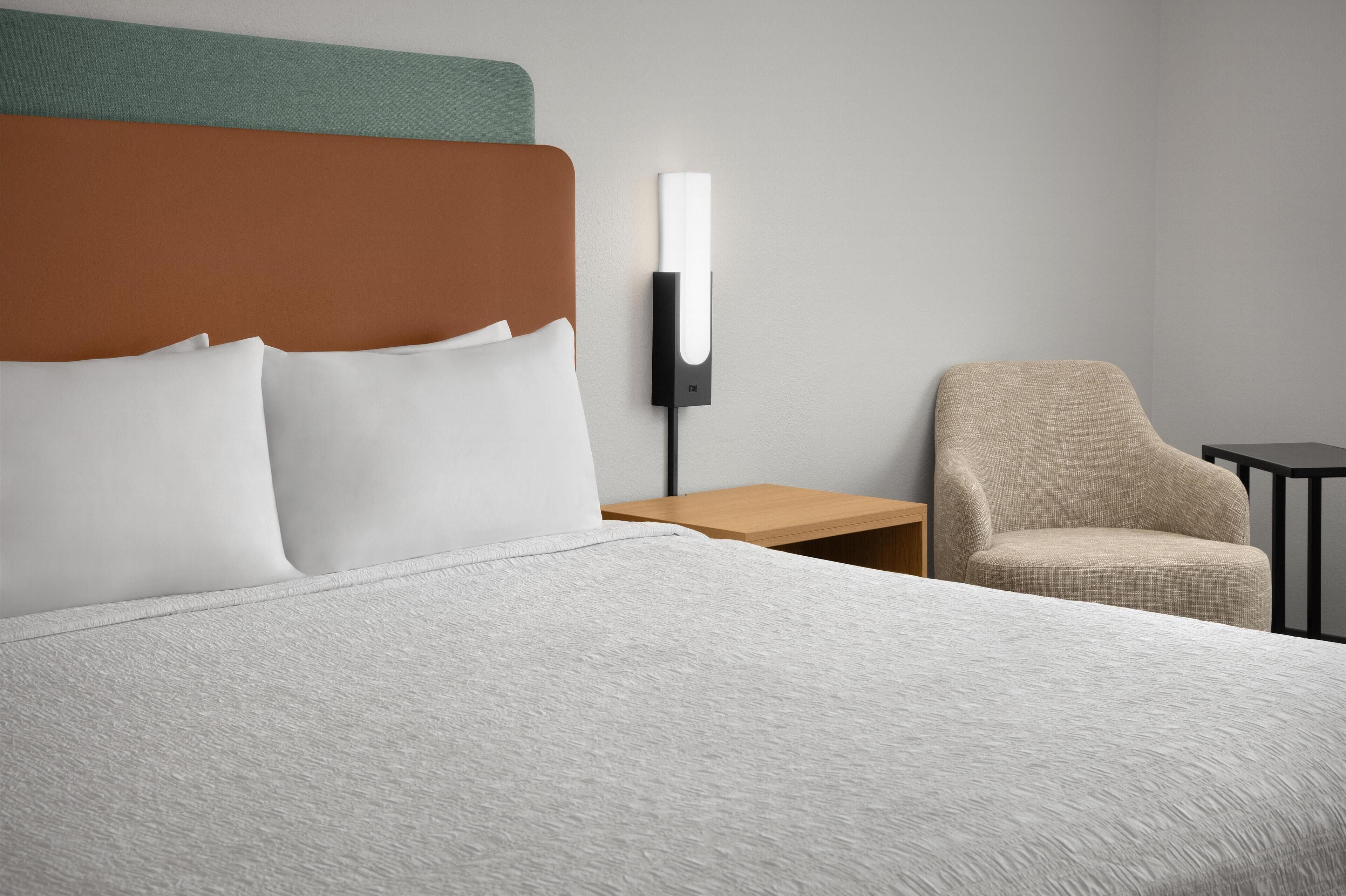 A hotel room with a bed, a nightstand, a lamp, a chair, and a small table.