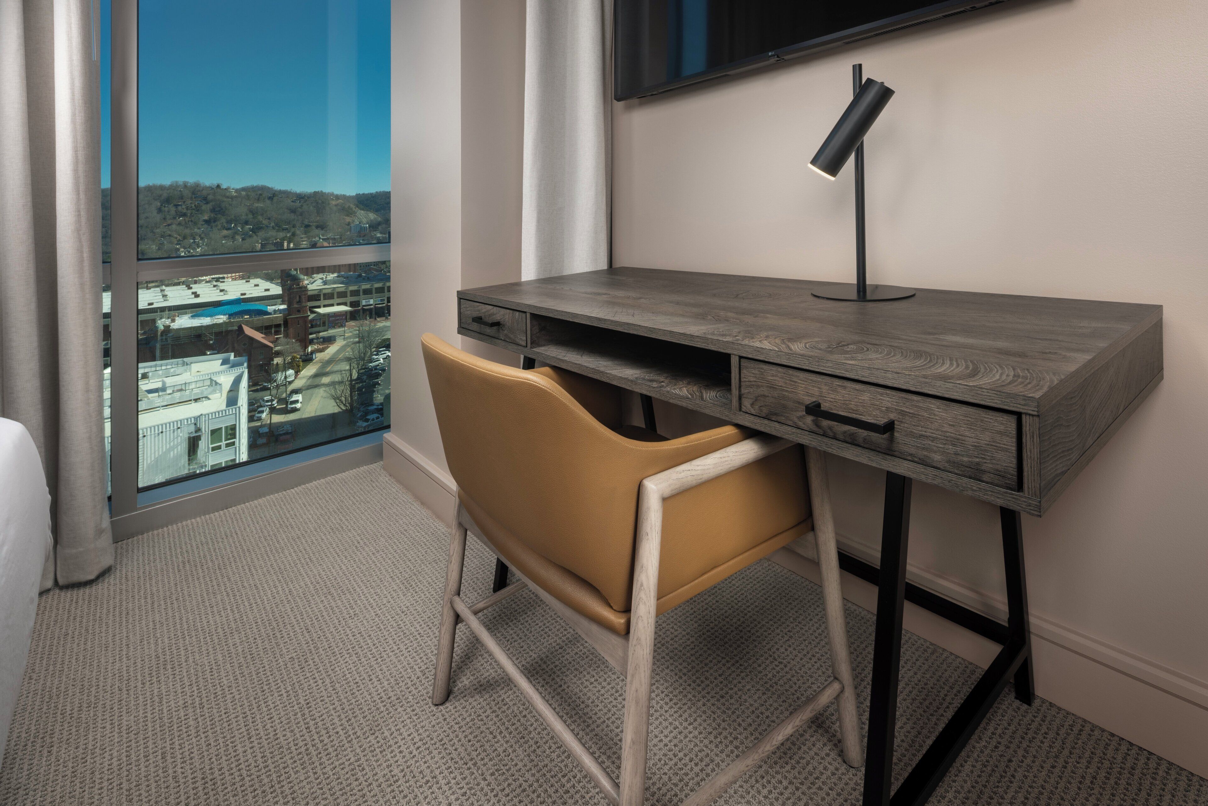 A modern hotel room with a large window offering a city view, a desk with a lamp, and a comfortable chair.