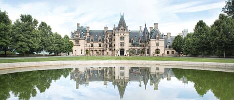 A large, historic building with a symmetrical facade and a central tower, surrounded by a reflective pond and trees.
