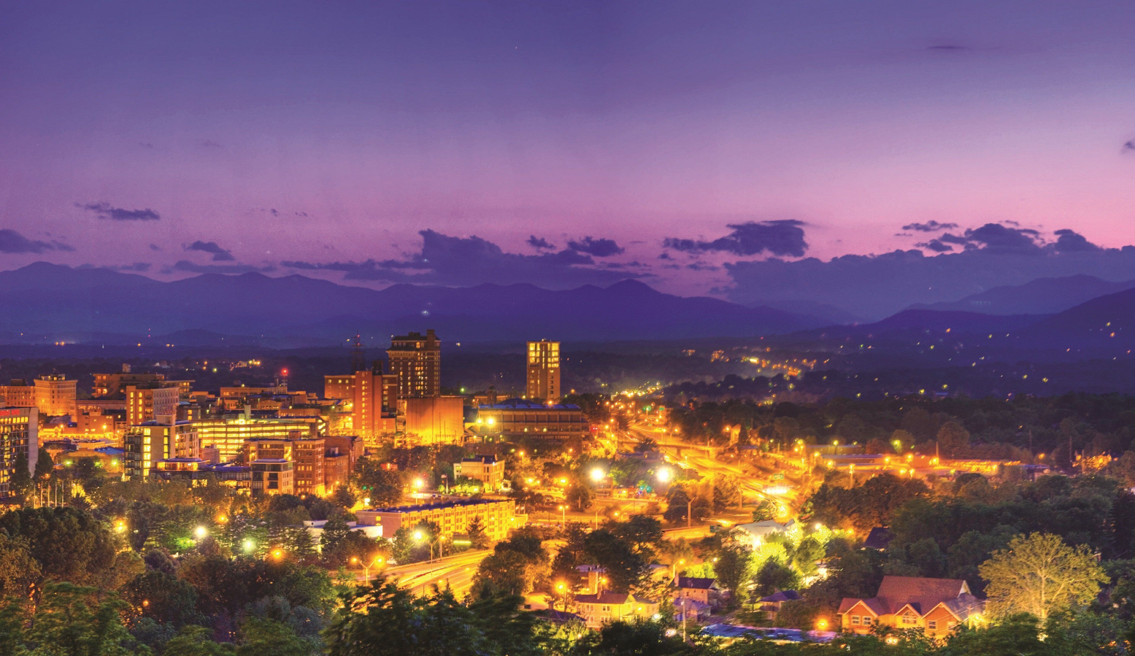 Une vue d'une ville au crépuscule, avec des bâtiments illuminés et une chaîne de montagnes en arrière-plan.