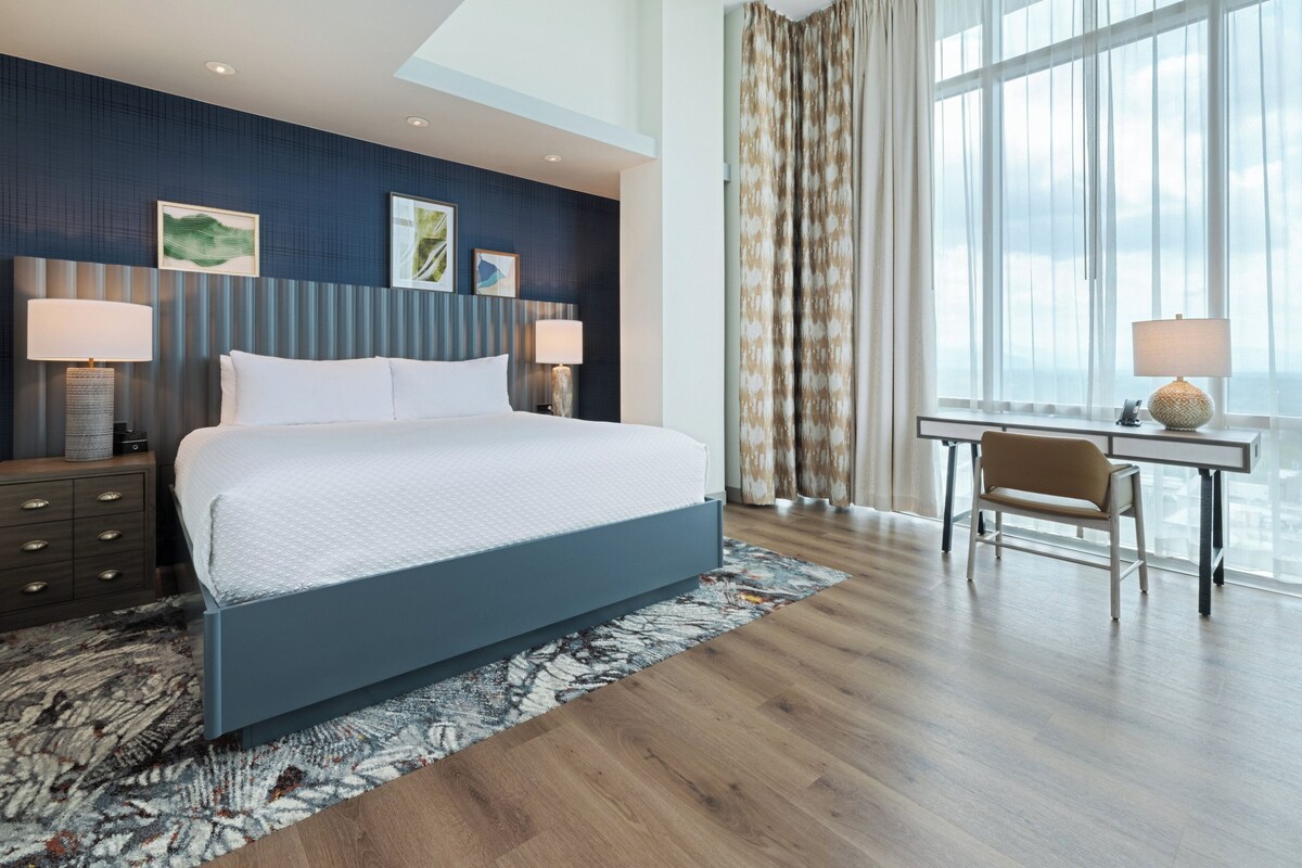 A modern hotel room with a large bed, two bedside tables with lamps, a desk with a chair, and a large window with curtains.