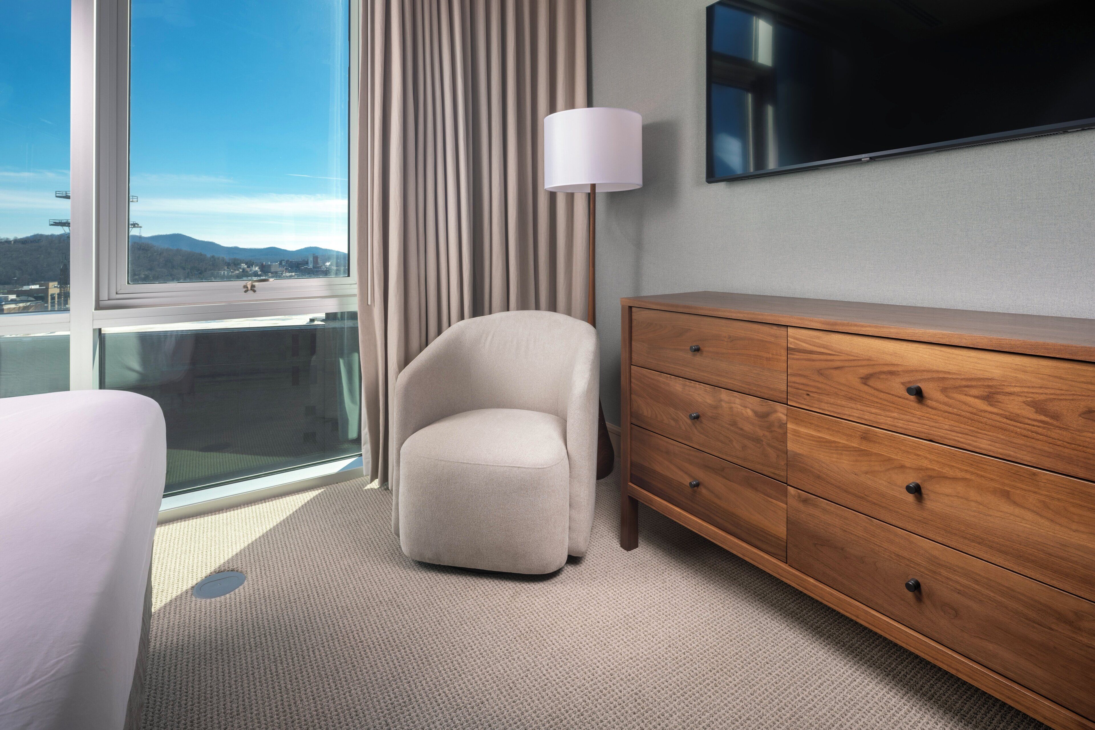 Habitación de hotel con un ventanal grande, un aparador de madera, una silla y una lámpara.