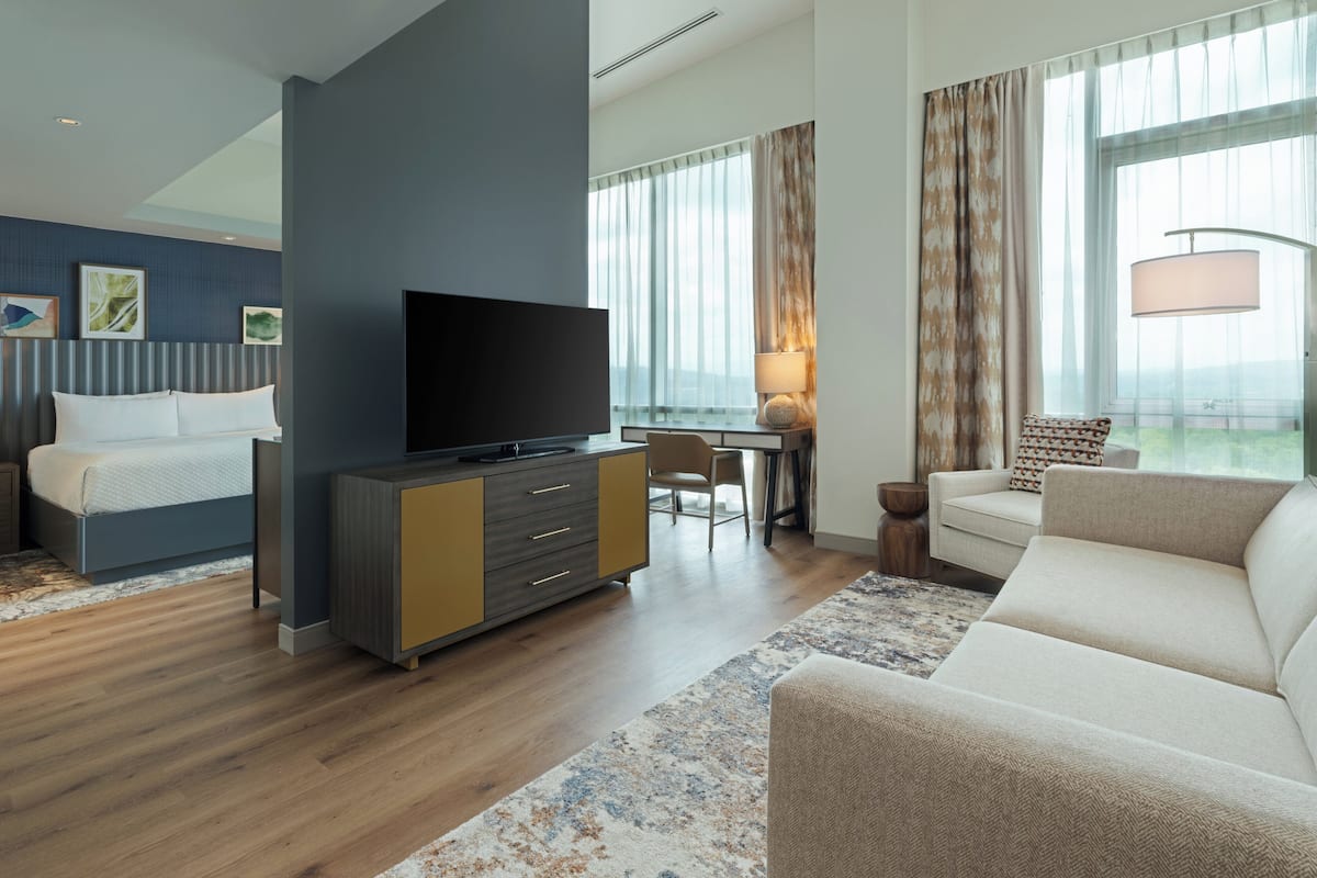A modern hotel room with a flat-screen TV, a sofa, a dining area, and large windows with curtains.