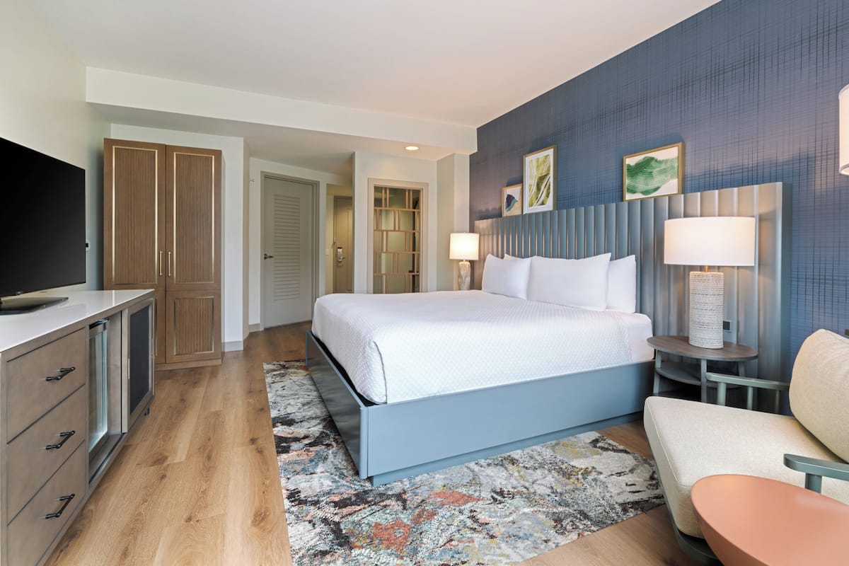 A modern hotel room with a large bed, a TV, wooden wardrobe, and a seating area with a table and chair.