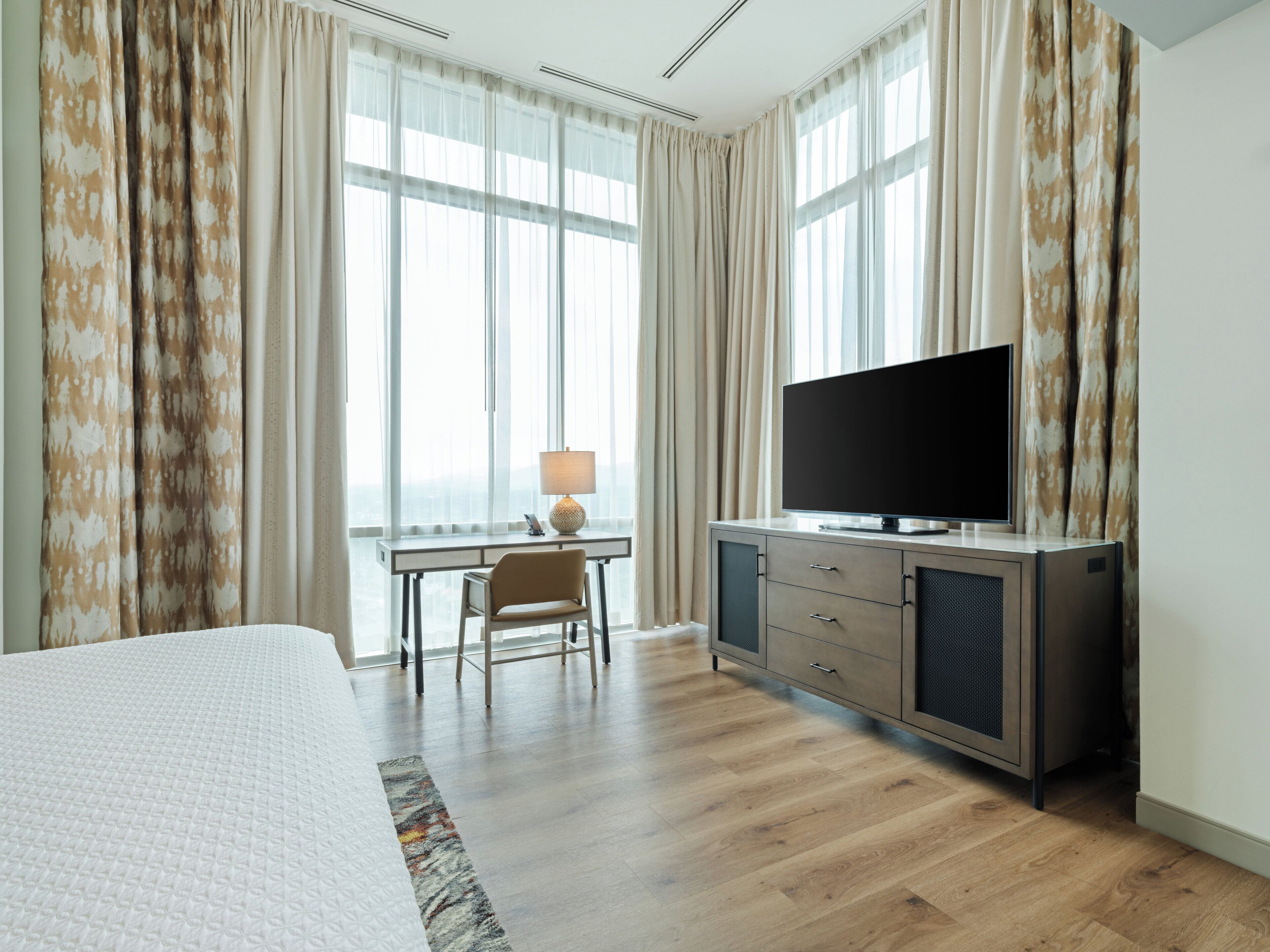 Quarto de hotel com cama, escrivaninha com luminária, televisão e vista da cidade através de janelas amplas.