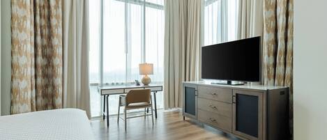 Quarto de hotel com cama, escrivaninha com luminária, televisão e vista da cidade através de janelas amplas.