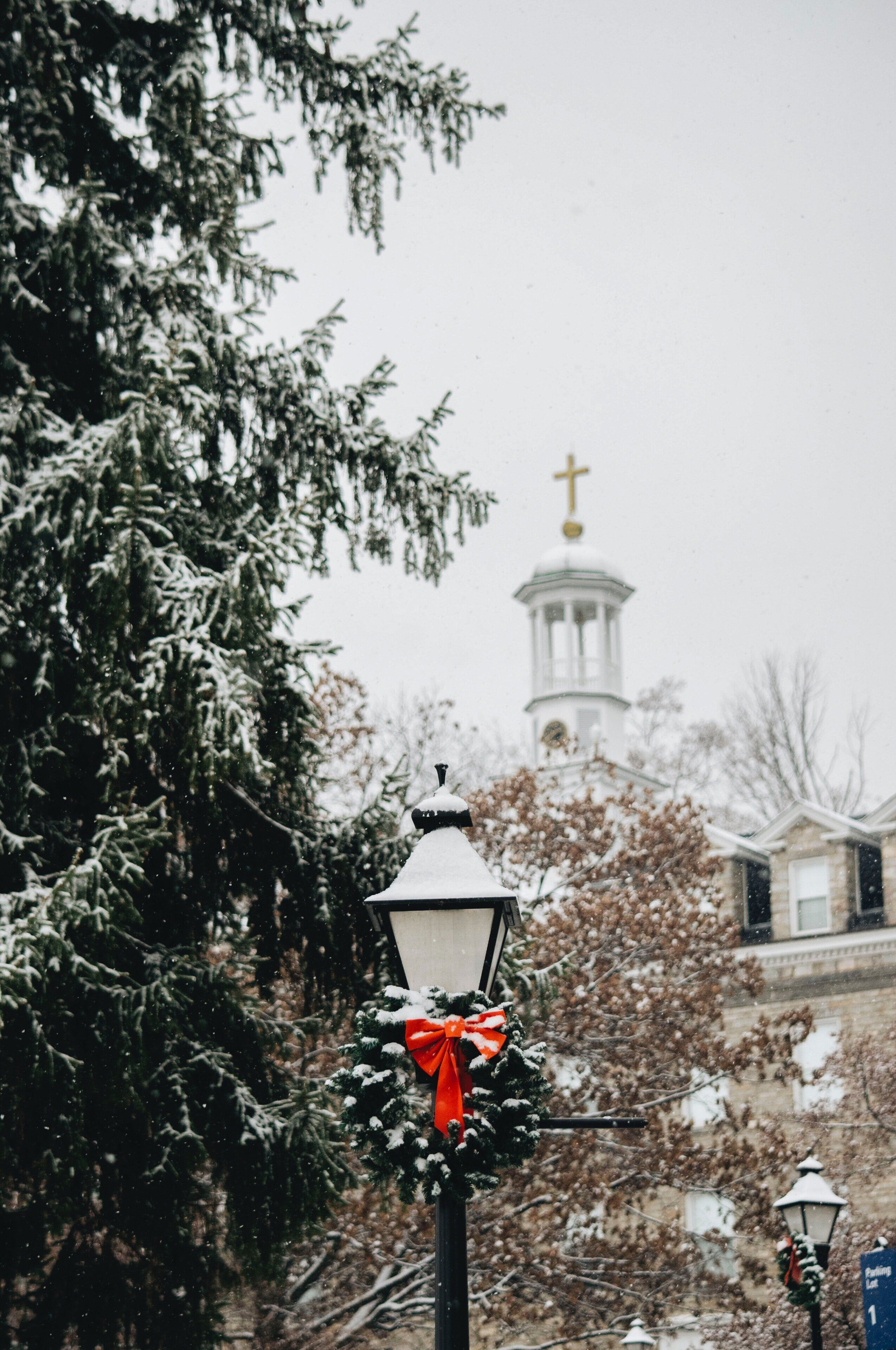 A snow-covered street lamp with a red bow, a church with a cross, and a building in the background.