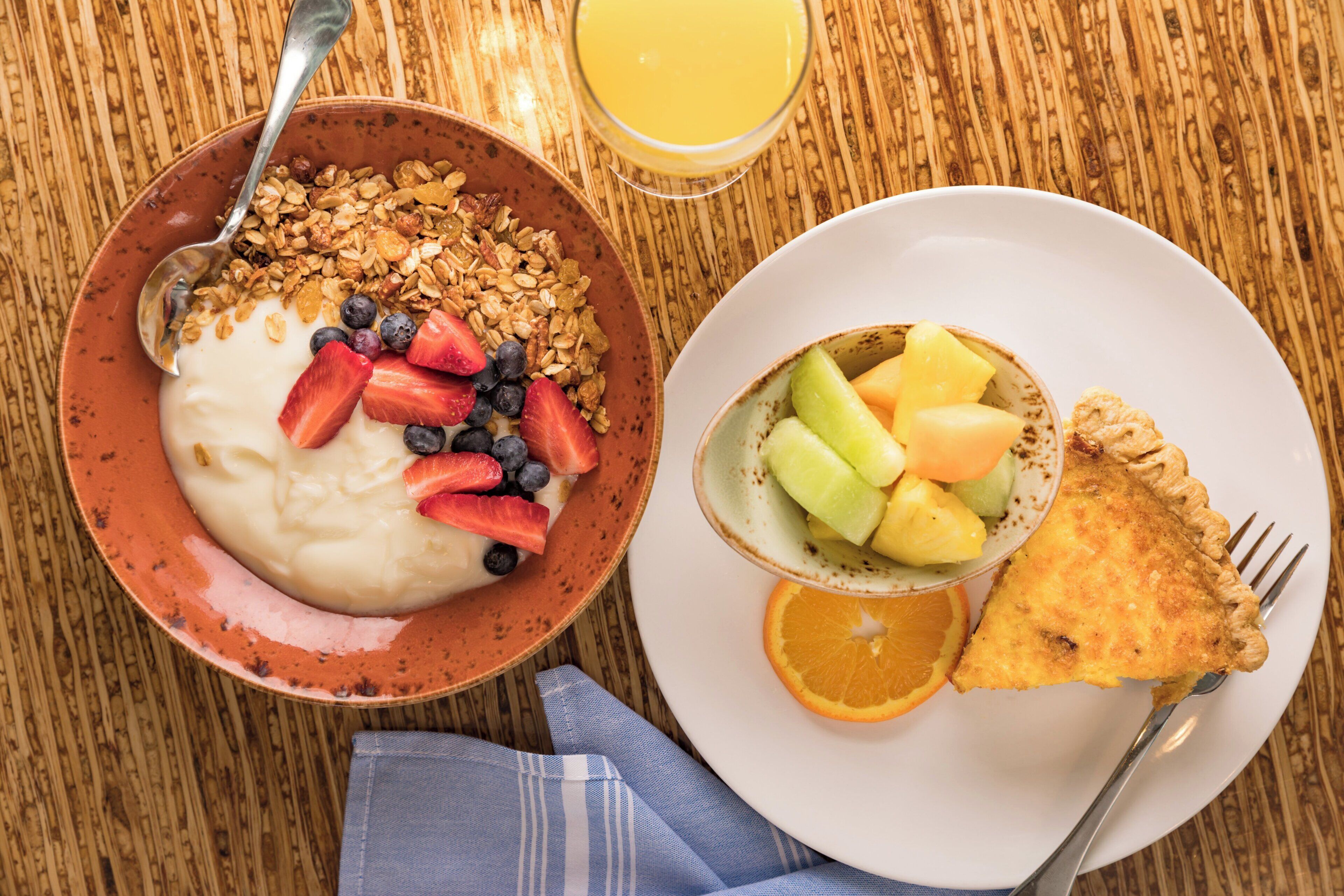 Een ontbijttafel met een kom yoghurt, belegd met fruit en granola, een bord met fruitsalade en een sneetje toast, en een glas sinaasappelsap op een houten tafel.