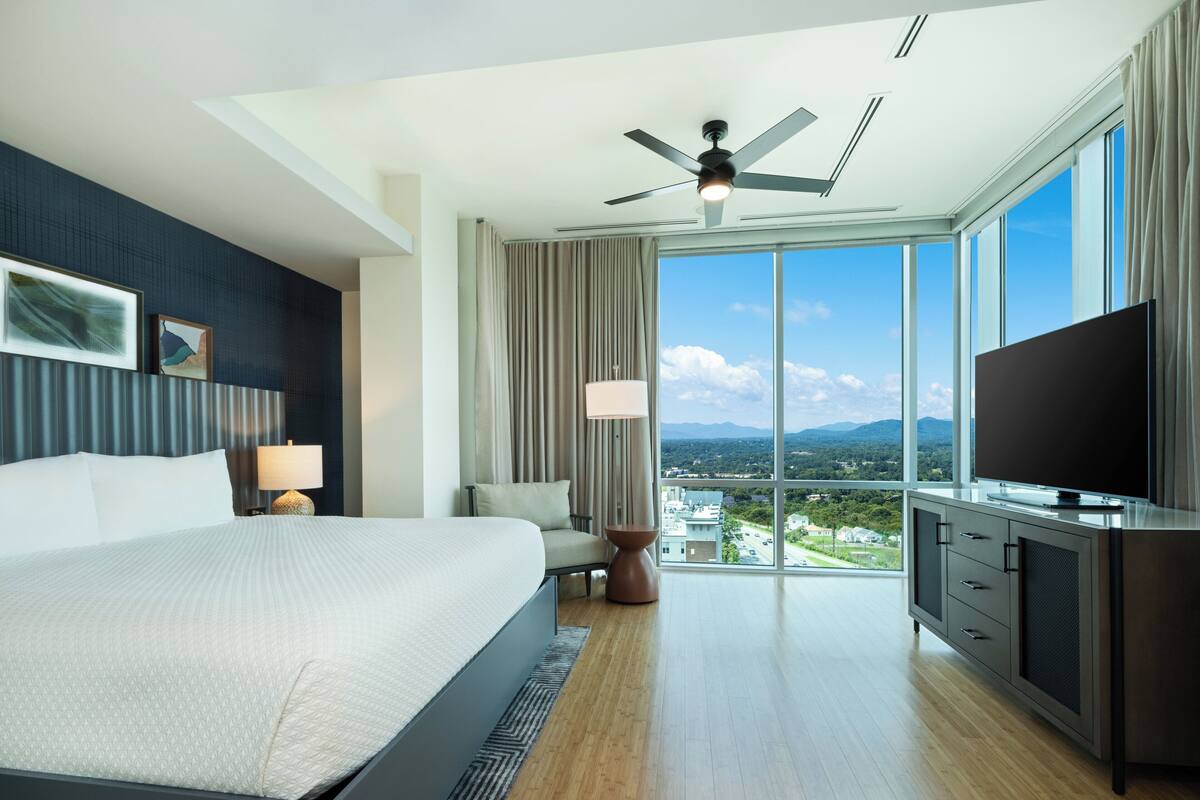 A modern hotel room with a large bed, a flat-screen TV, a ceiling fan, and a view of a cityscape through large windows.