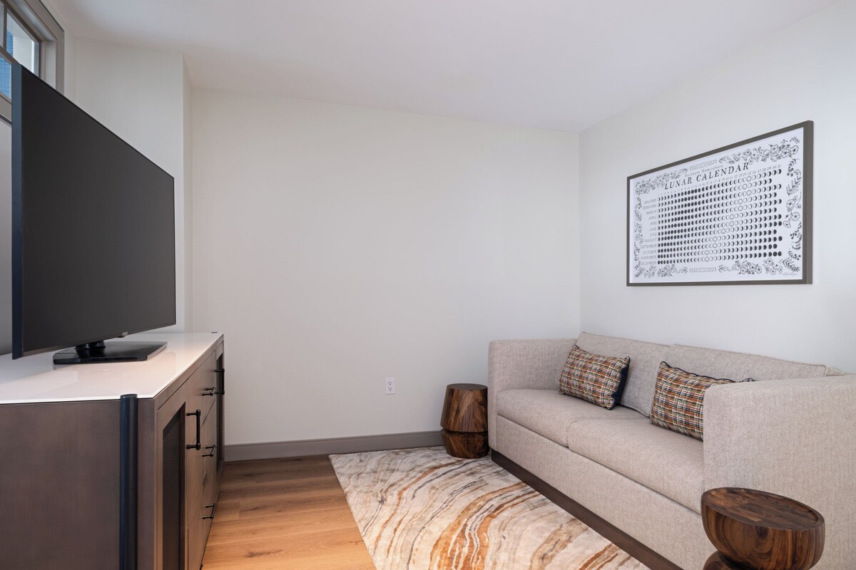 A modern living room with a flat-screen TV, a sofa, a patterned rug, and a wall-mounted calendar.