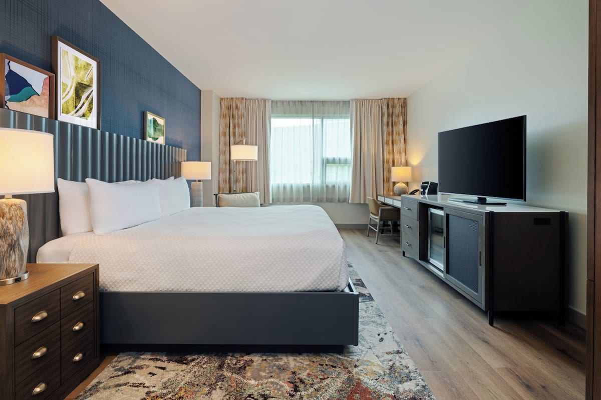 A modern hotel room with a large bed, a desk with a flat-screen TV, and a seating area with a chair and lamp.