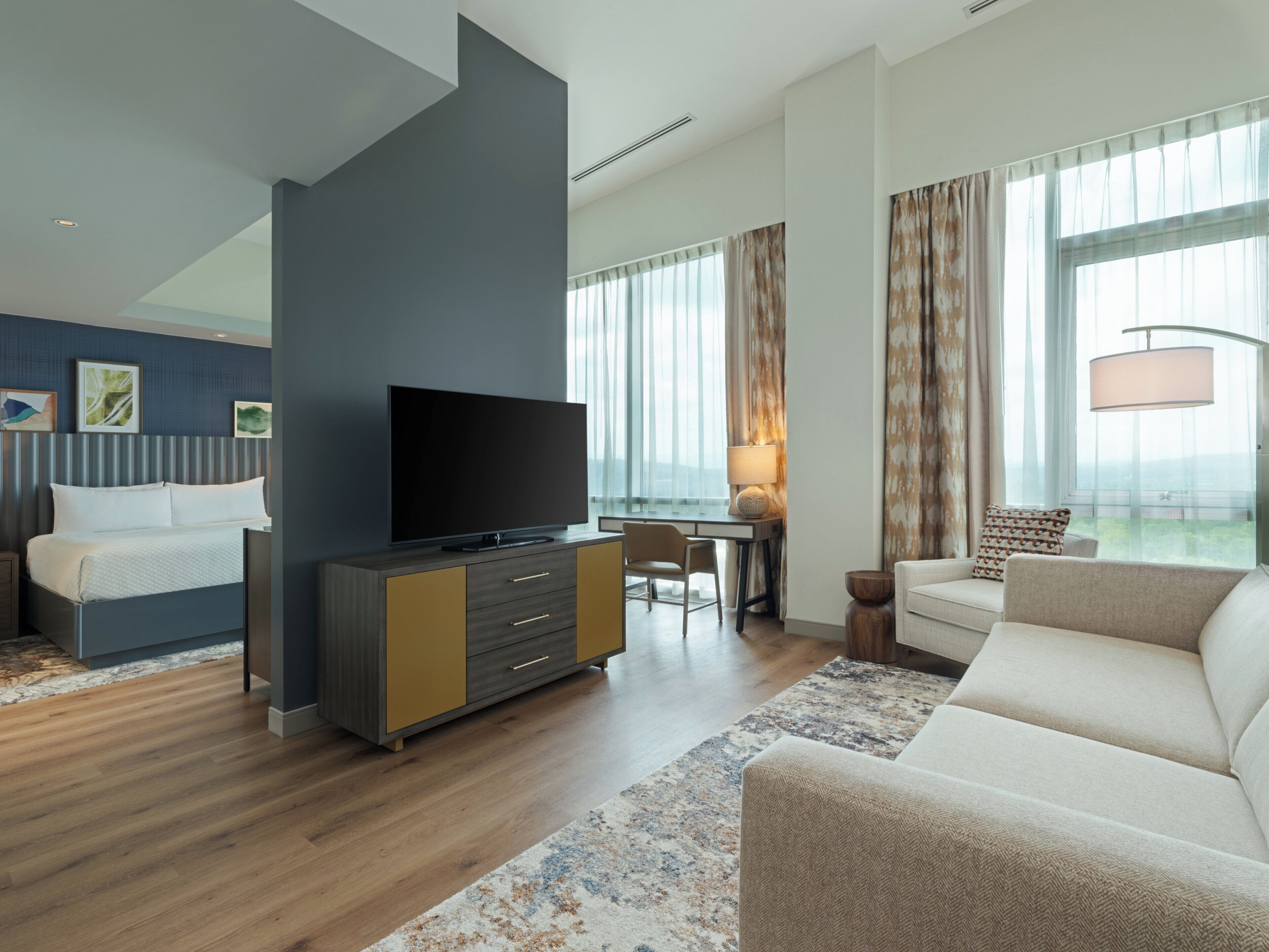 A modern hotel room with a flat-screen TV, a sofa, a dining area, and large windows with curtains.