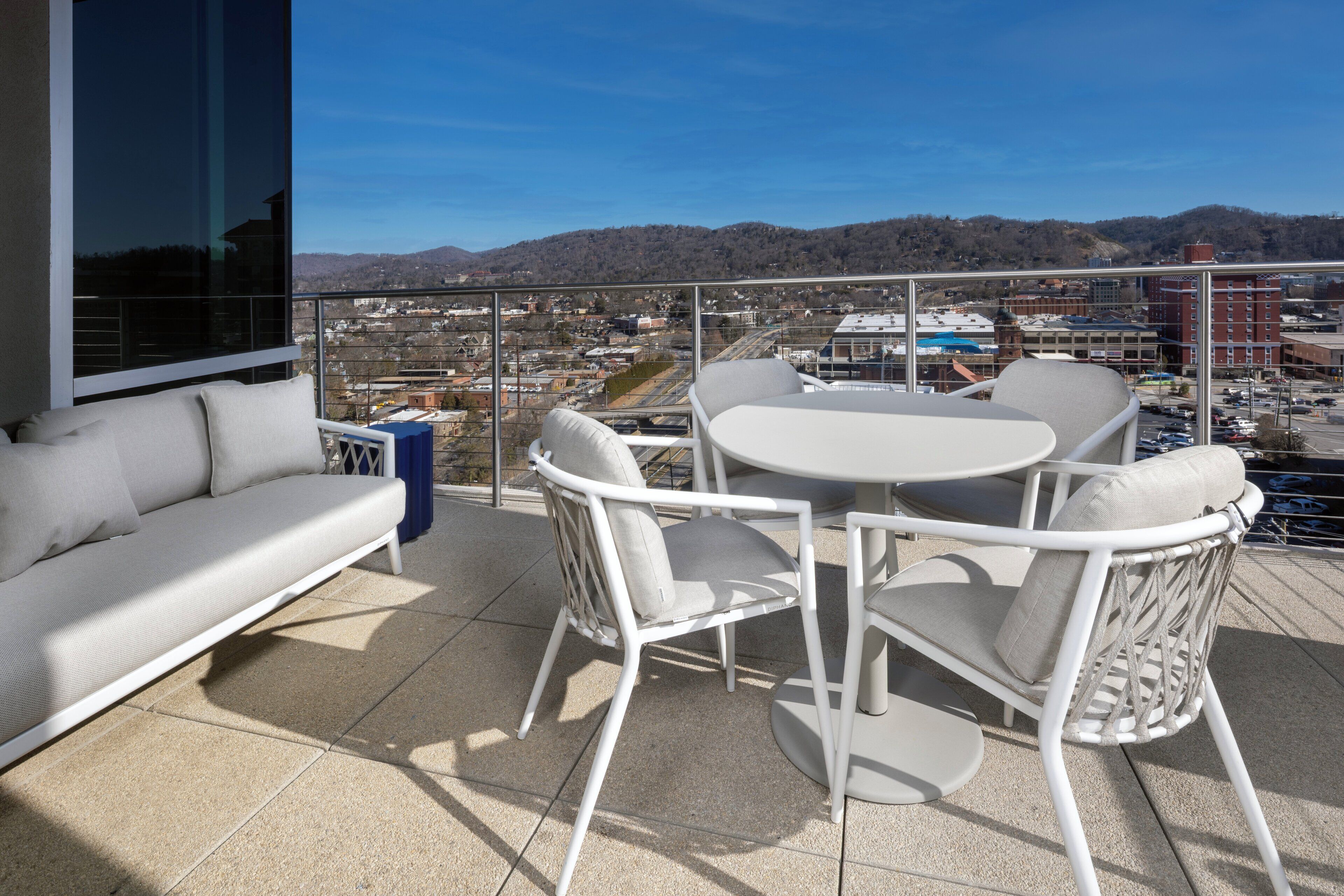 En balkon med en hvid sofa og stole, et rundt bord samt udsigt til by og bjerge.