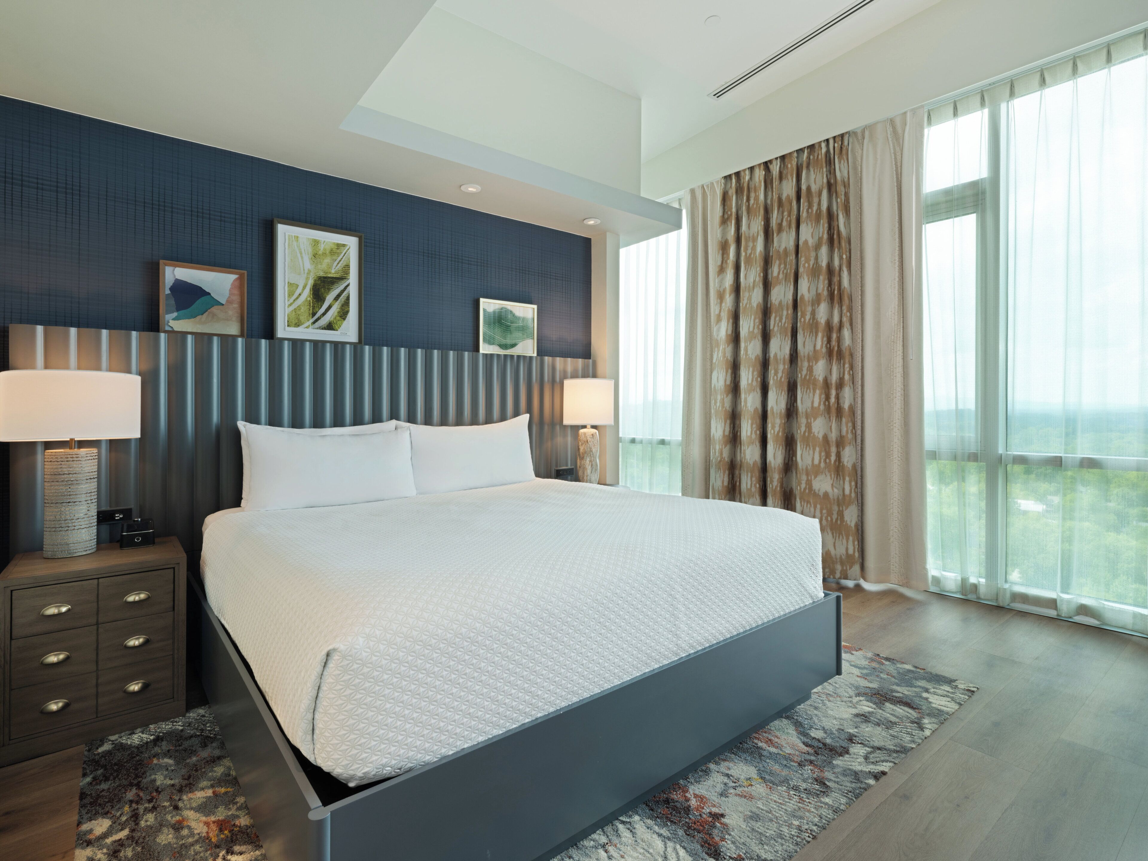 Une chambre d’hôtel moderne avec un grand lit, deux tables de chevet avec des lampes, un tapis à motifs et une vue sur la ville à travers de grandes fenêtres.