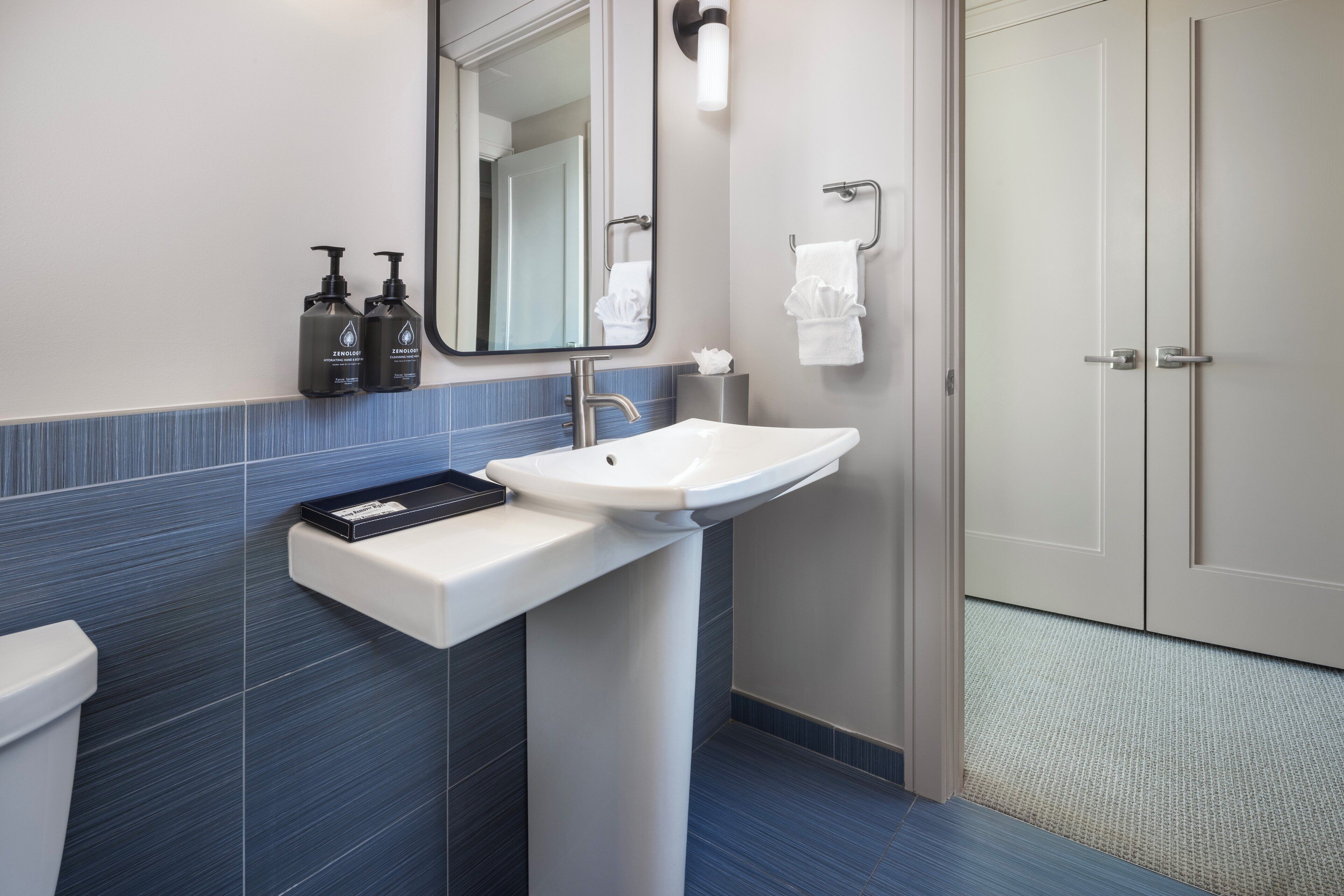 A modern bathroom with a white sink, a mirror, and a towel rack. There are two bottles of shampoo and conditioner on the counter.