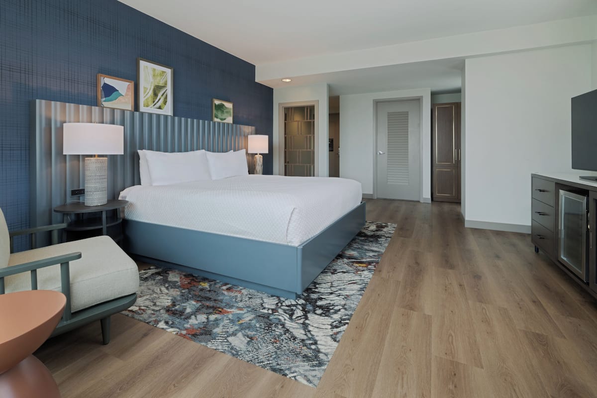 A modern hotel room with a large bed, bedside tables with lamps, a TV, and a seating area with a chair and a small table.