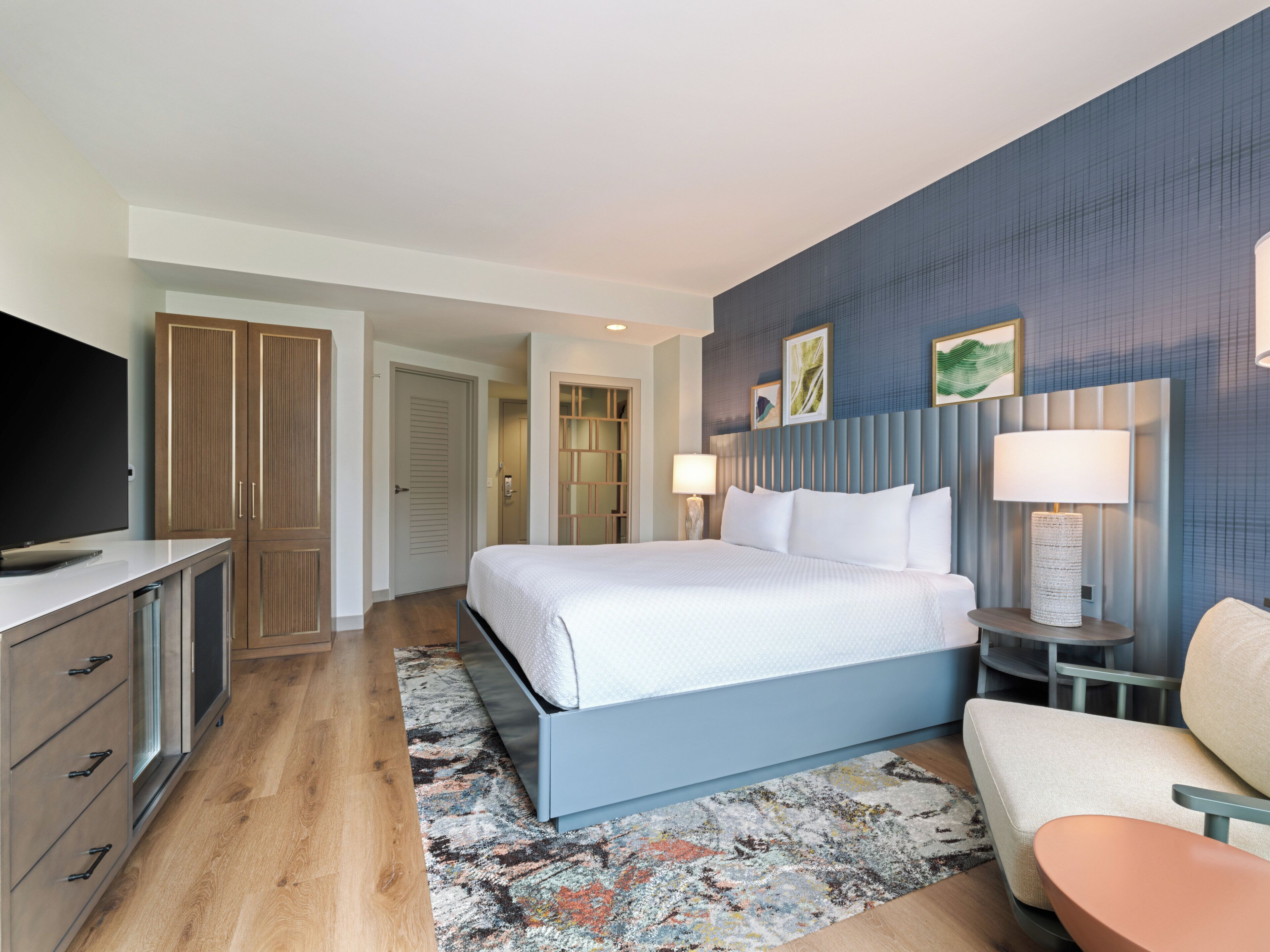 A modern hotel room with a large bed, a TV, wooden wardrobe, and a seating area with a table and chair.