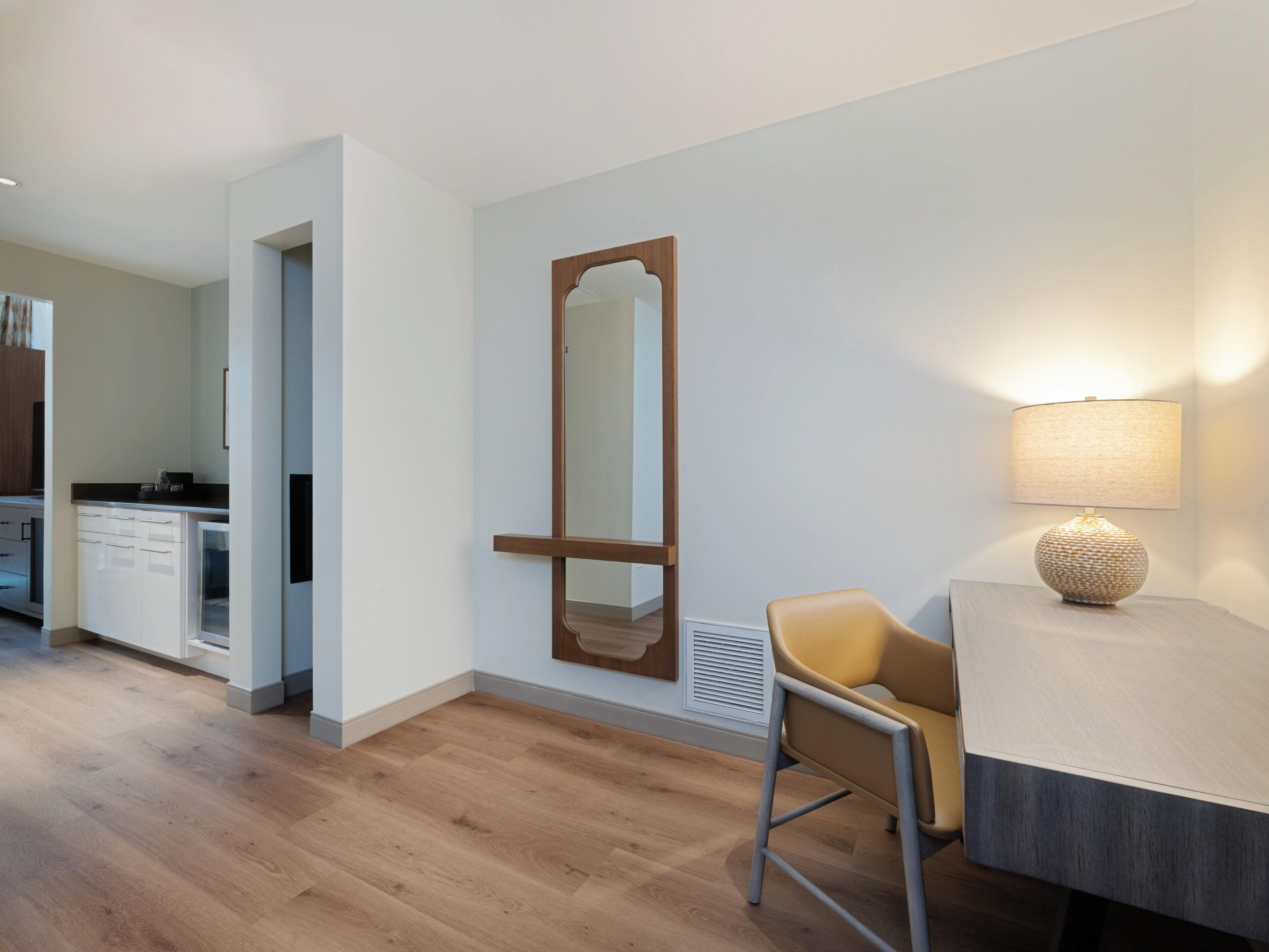 Una habitación moderna con suelo de madera, un escritorio con lámpara, una silla y un espejo grande en la pared.