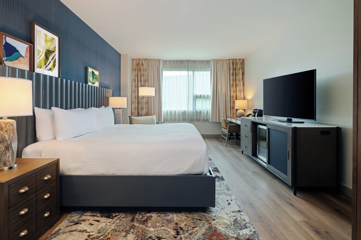 A modern hotel room with a large bed, a desk with a flat-screen TV, and a seating area with a chair and lamp.