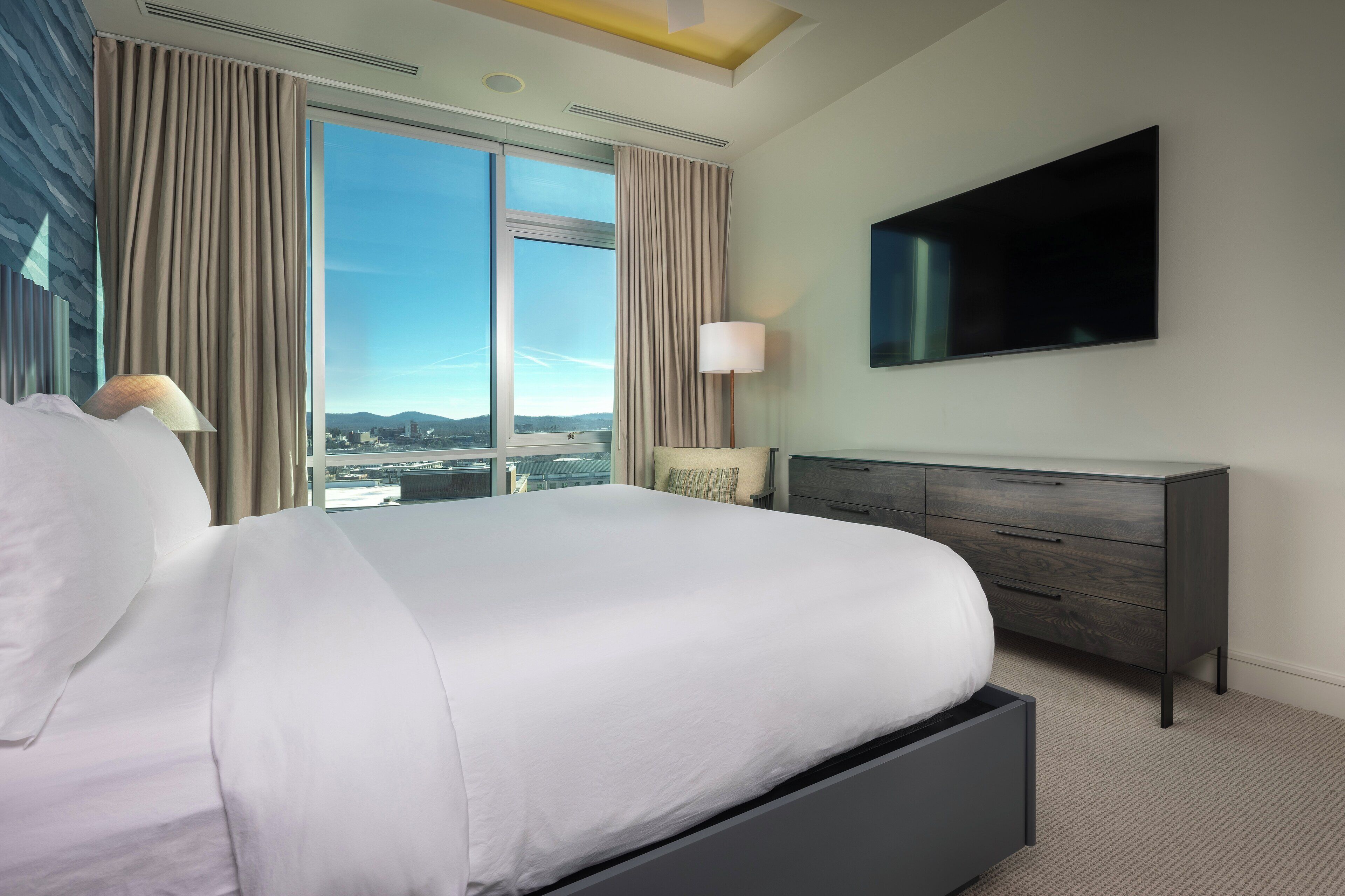 Una habitación de hotel moderna con una cama grande, un televisor de pantalla plana, una mesita de noche y vistas a un paisaje urbano a través de ventanales de techo a piso.
