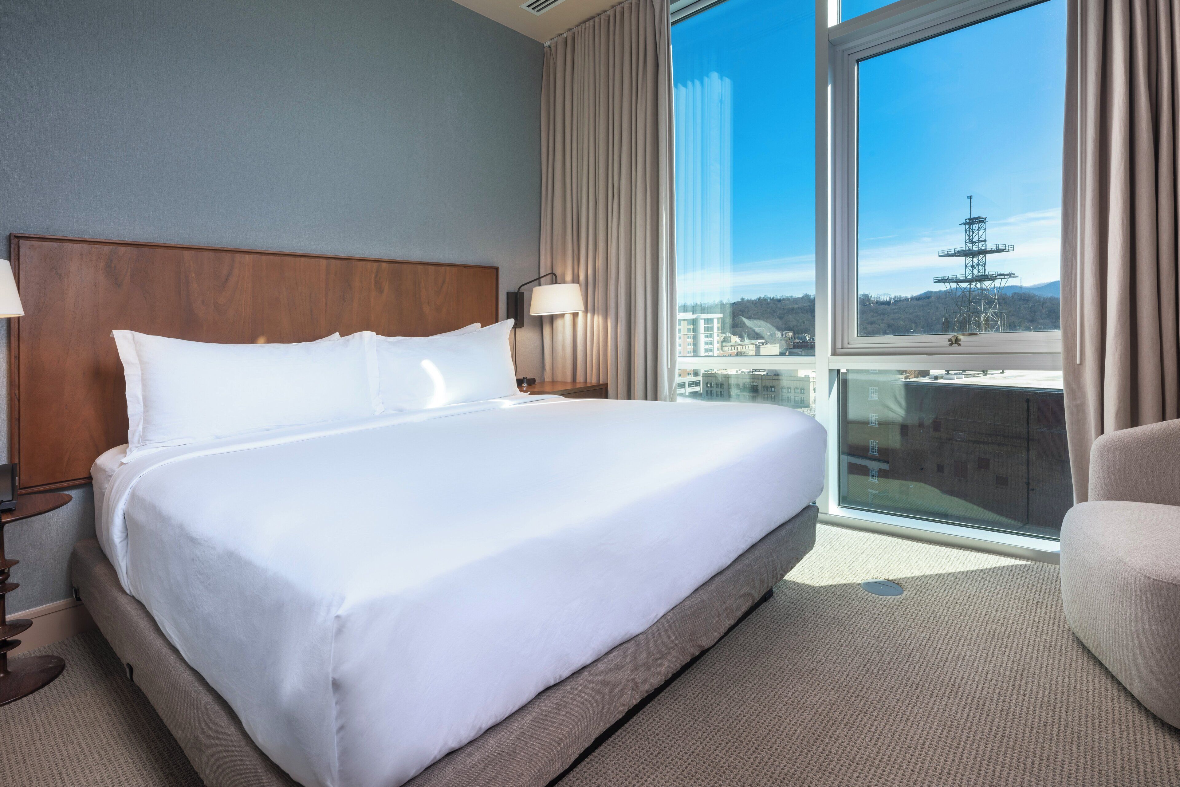 Une chambre d’hôtel avec un grand lit, une table de chevet, une lampe et une vue sur le paysage urbain, visible à travers une grande fenêtre.