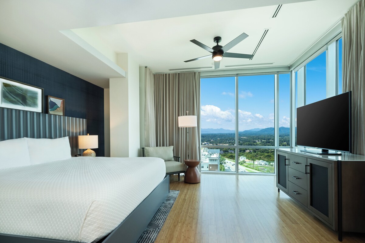 A modern hotel room with a large bed, a flat-screen TV, a ceiling fan, and a view of a cityscape through large windows.